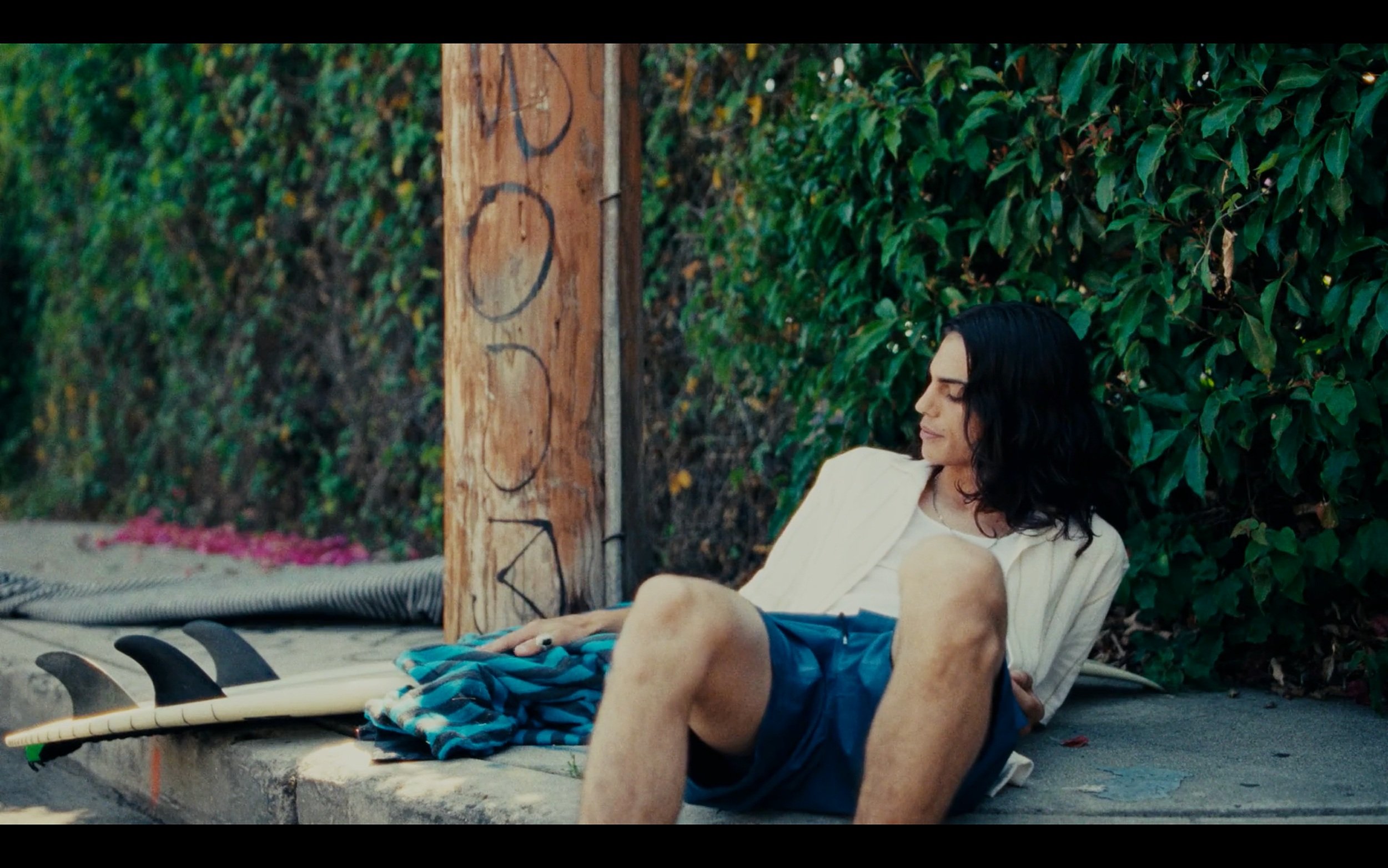 Un homme aux cheveux longs assis sur un trottoir, adossé à un mur recouvert de feuilles. À côté de lui, il y a une planche de surf et une serviette bleue.