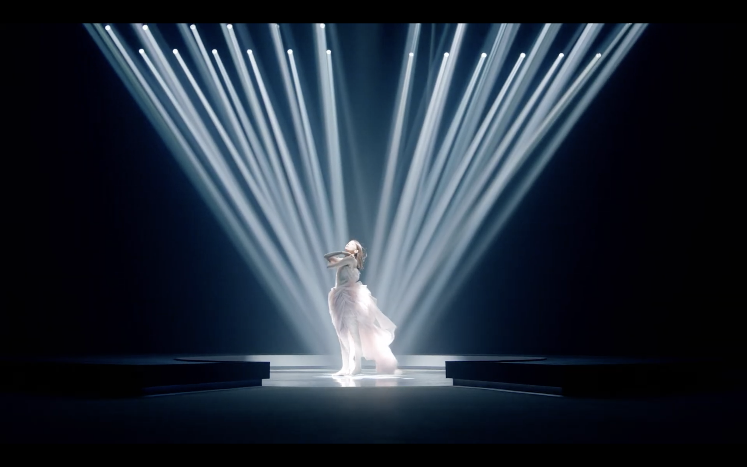 Une personne en robe élégante debout sur scène, illuminée par des projecteurs convergents.