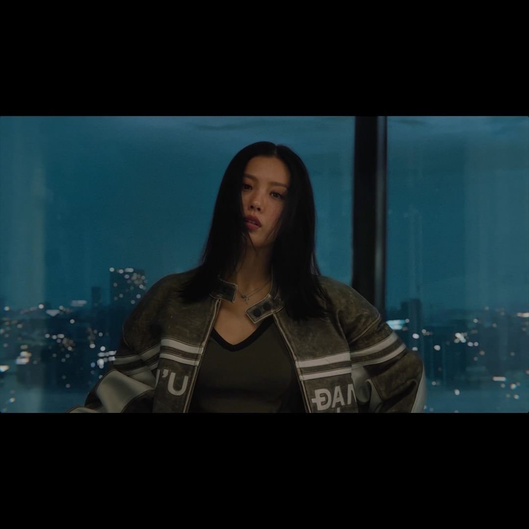 Femme aux cheveux noirs, portant un blouson, devant une fenêtre donnant sur une vue nocturne de la ville.