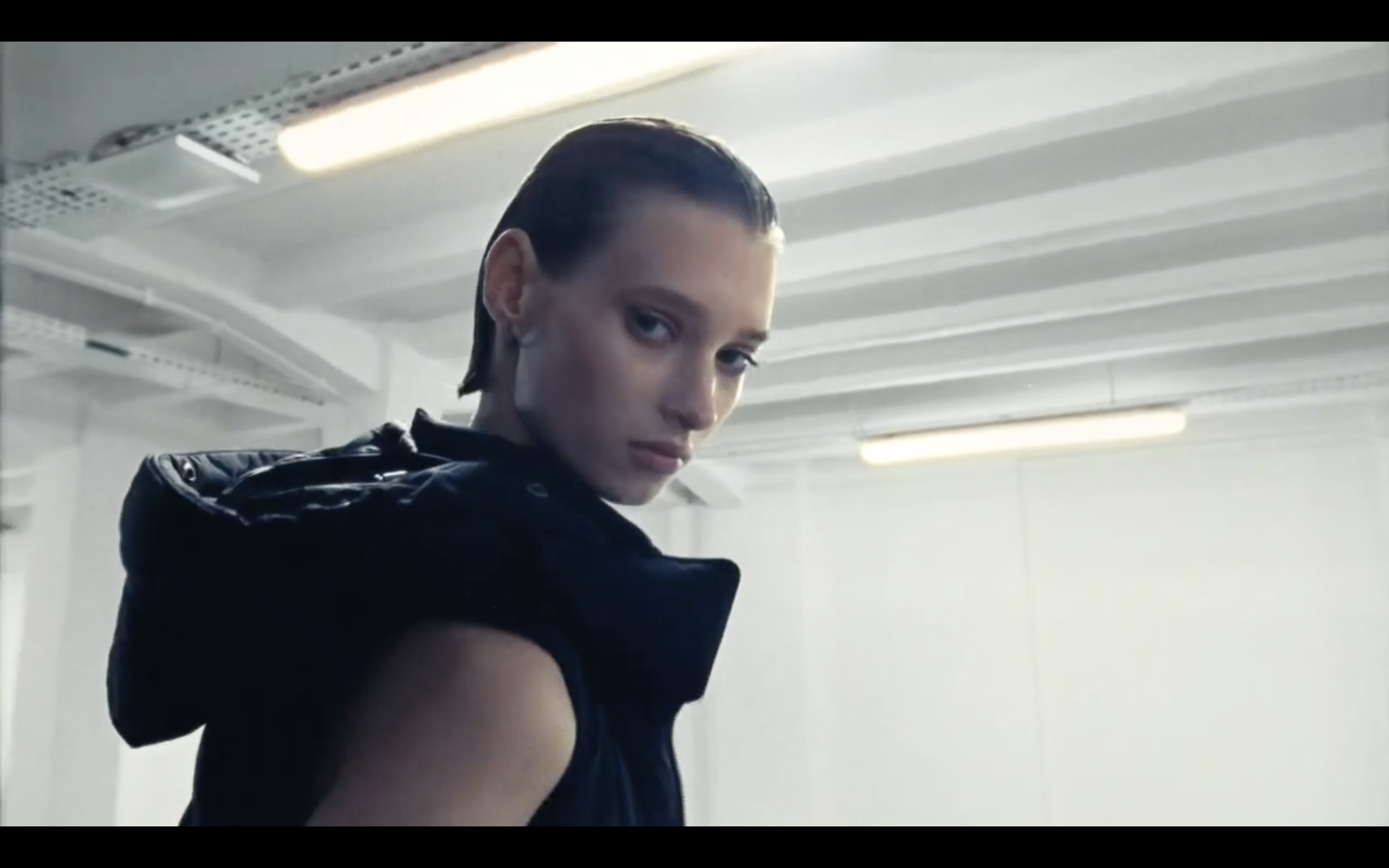 Personne regardant par-dessus l'épaule, portant un manteau noir dans un environnement industriel lumineux.