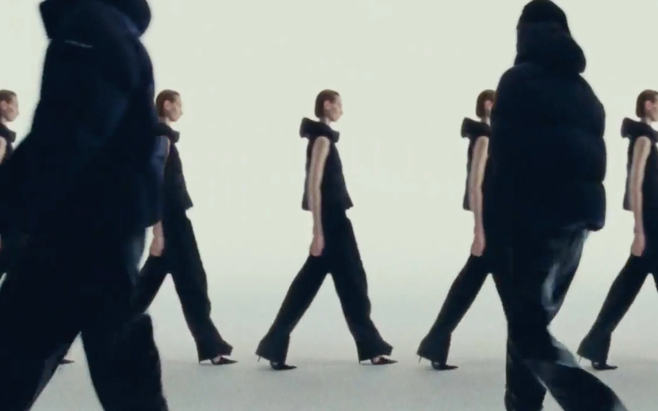 Des mannequins défilent en vêtements noirs sur un fond clair.