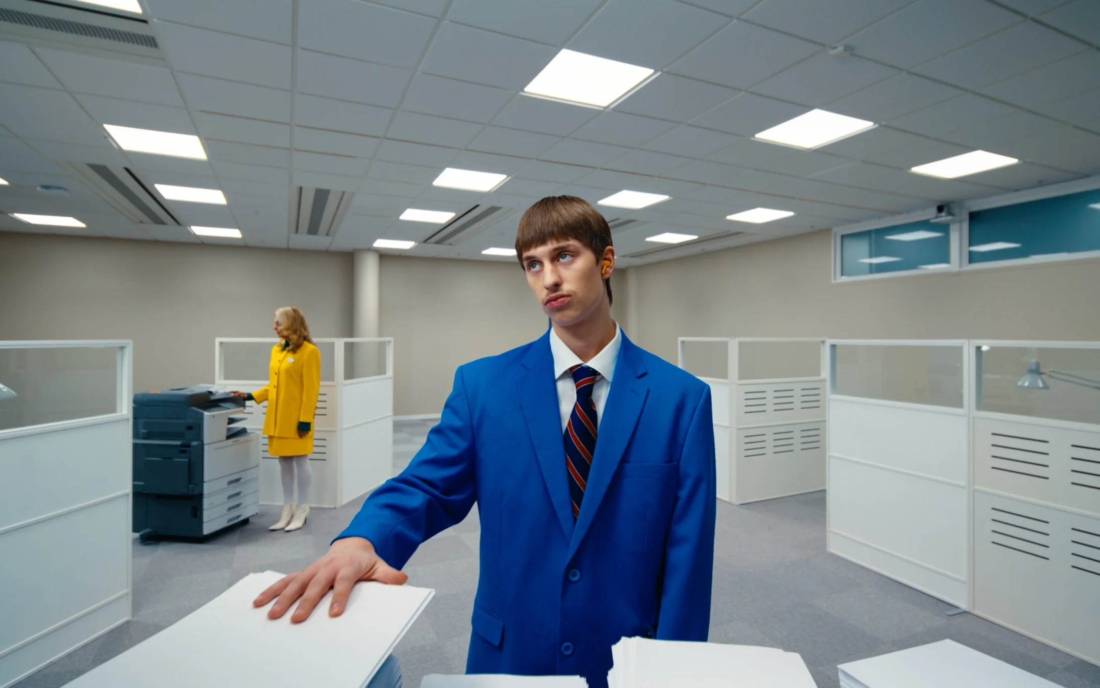 Deux personnes dans un bureau moderne, portant des costumes colorés, travaillent près d'une photocopieuse et des papiers, dans un espace de travail ouvert.