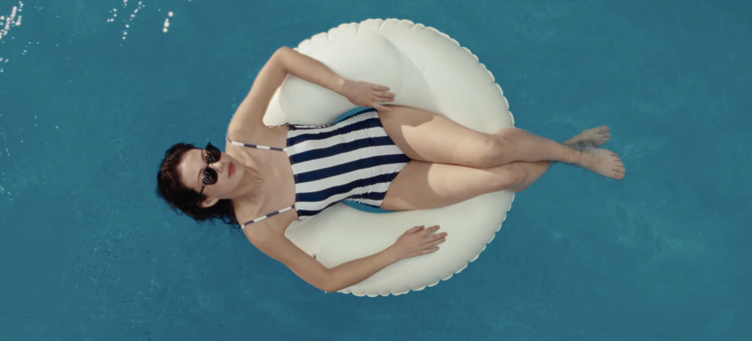 Femme flottant dans une piscine sur une bouée blanche, portant un maillot de bain rayé et des lunettes de soleil.