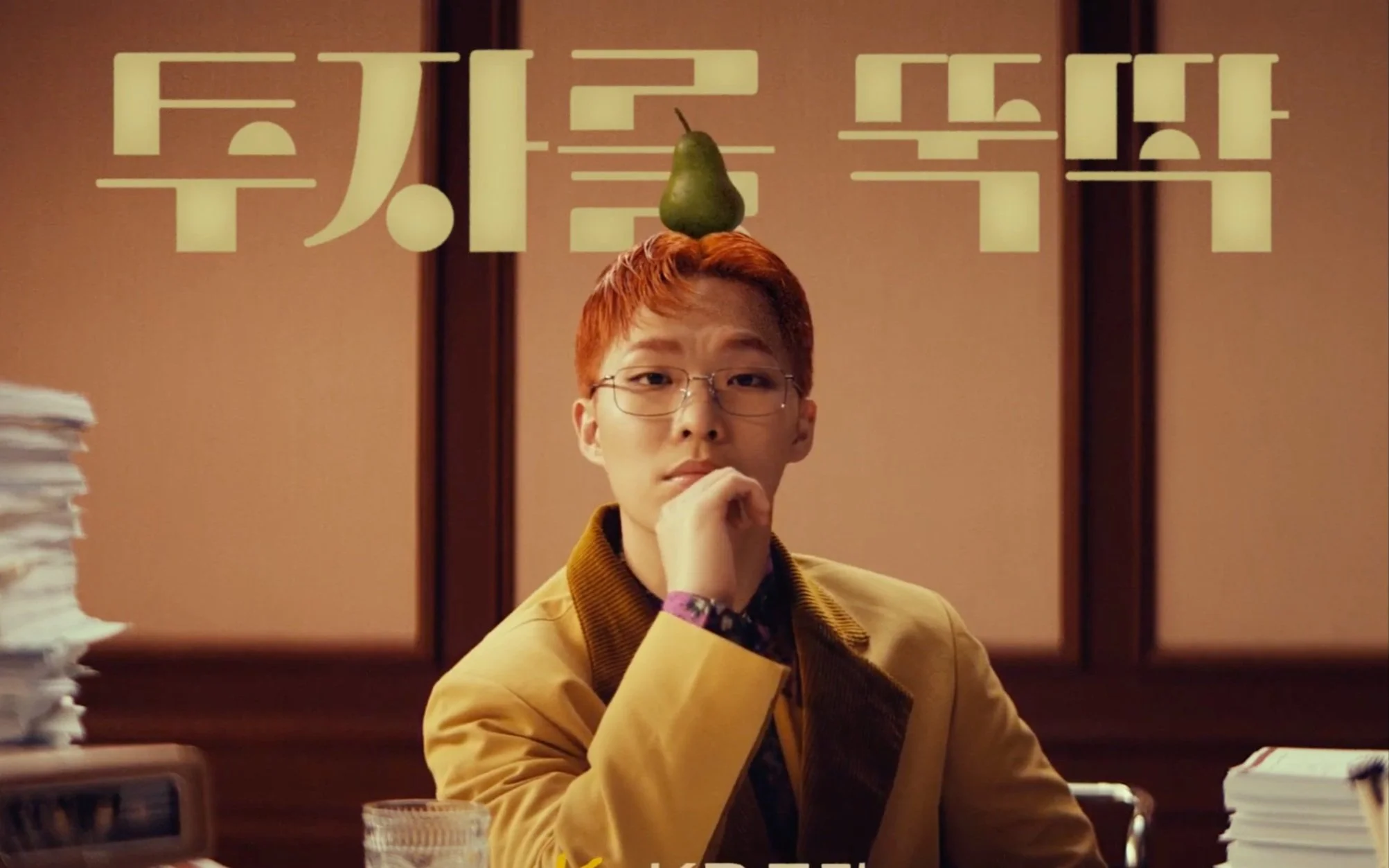 Personne assise à un bureau avec une poire sur la tête, portant des lunettes et un costume jaune. Texte en caractères coréens en arrière-plan.