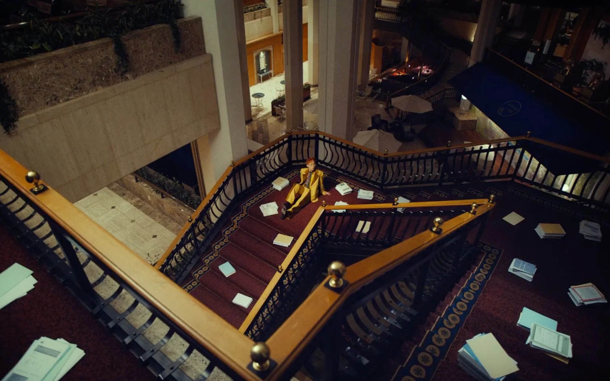 Intérieur d'un bâtiment avec un grand escalier, où une personne en costume jaune est assise au milieu de papiers éparpillés.