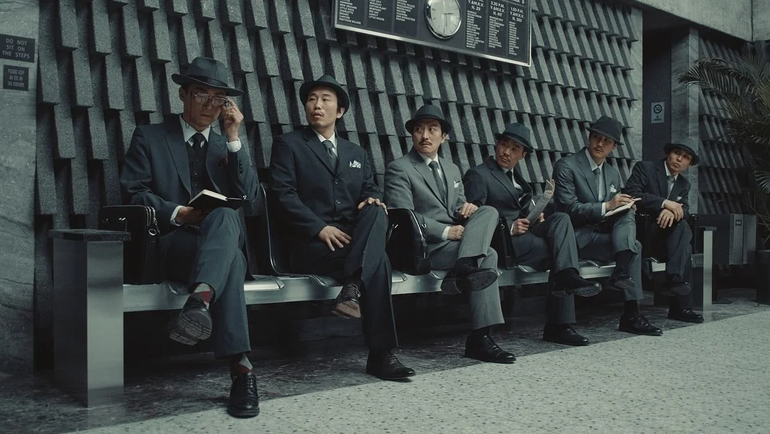 Six hommes en costumes et chapeaux sont assis sur un banc dans un espace public, avec un panneau d'affichage et une horloge au mur.
