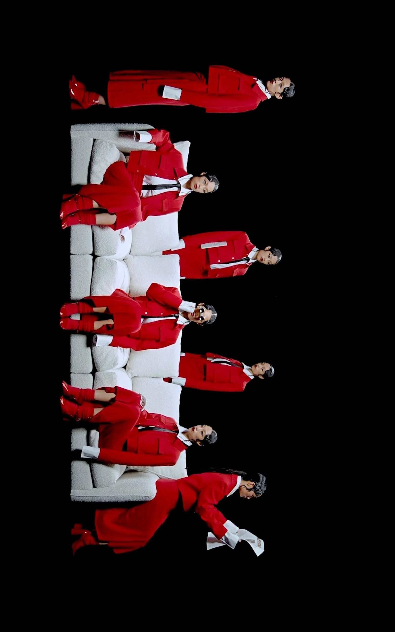 Plusieurs personnes en costumes rouges assises et debout autour d'un canapé blanc, fond noir.