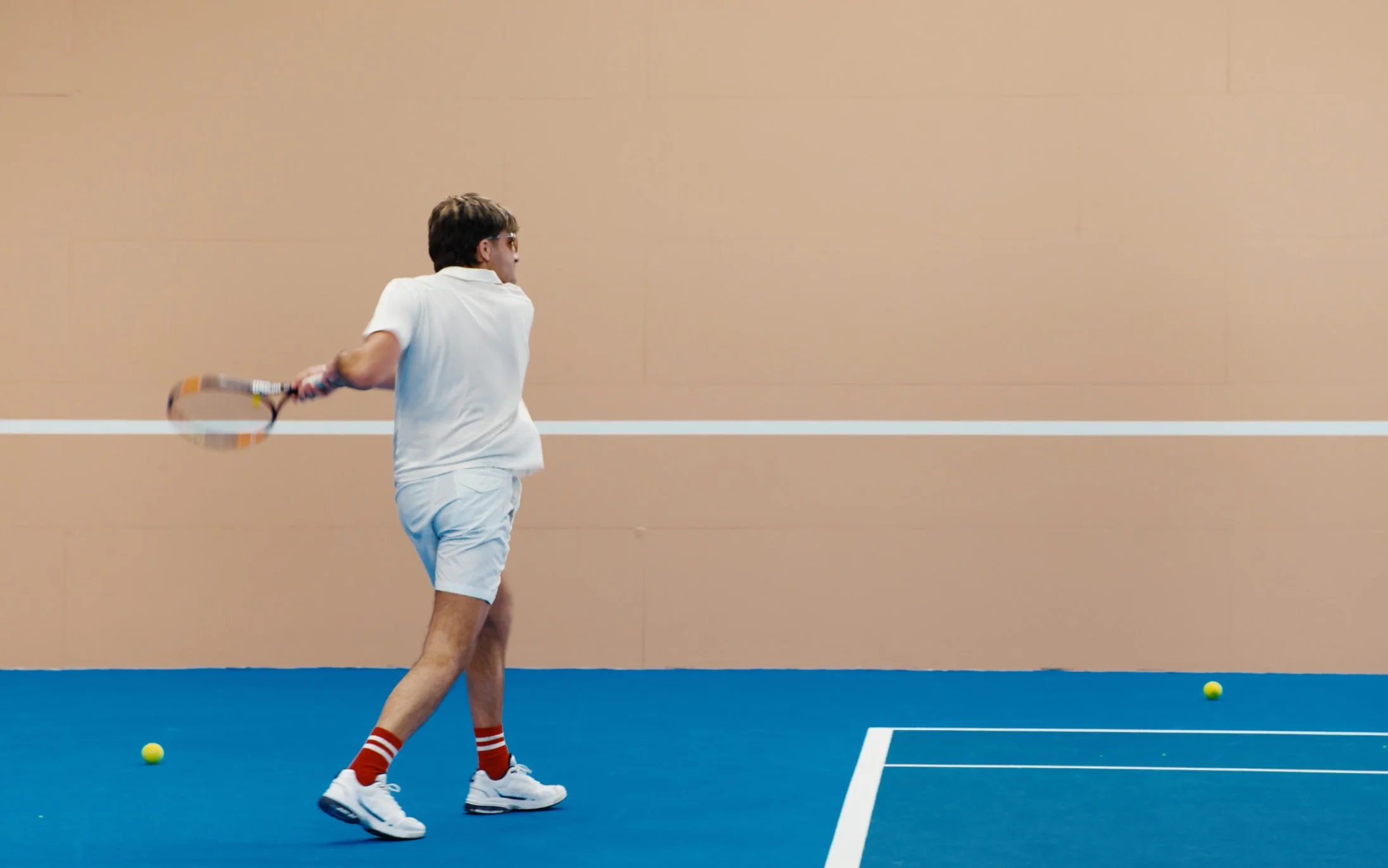 Un homme joue au tennis, vêtu de blanc, frappant une balle sur un terrain intérieur.