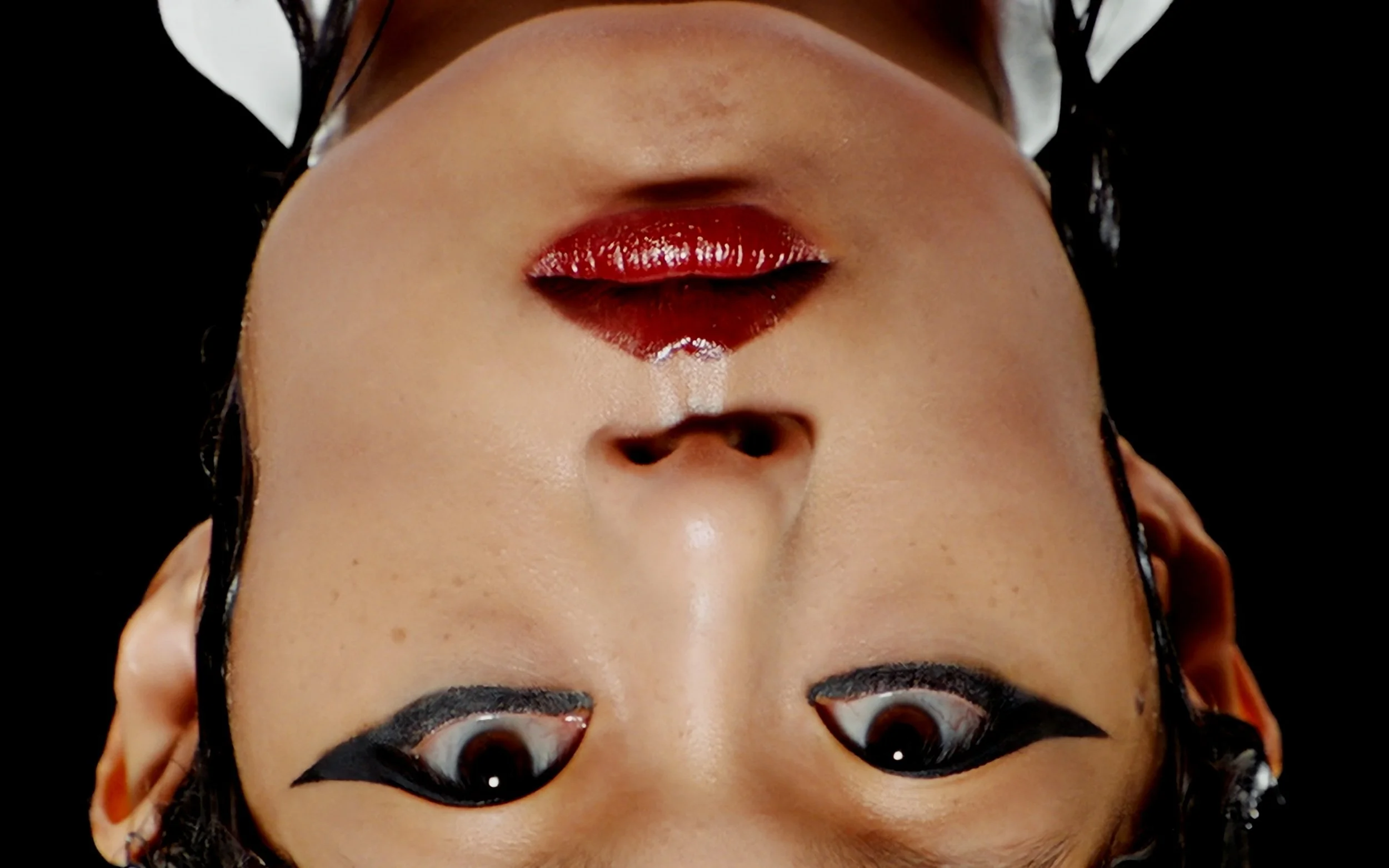 Visage d'une personne maquillée, à l'envers, avec des lèvres rouges brillantes et des yeux avec un maquillage accentué noir.