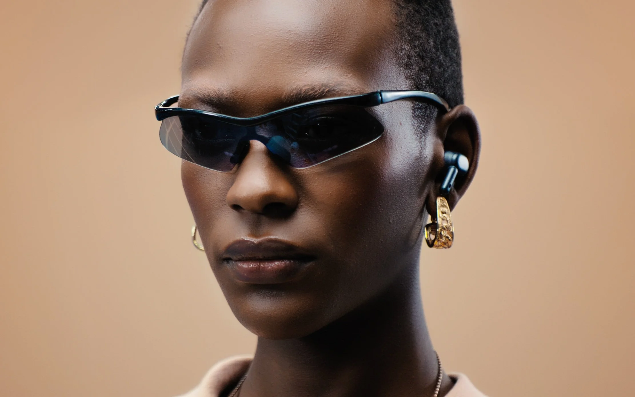 Une personne portant des lunettes de soleil futuristes et des boucles d'oreilles en or, sur un fond beige.
