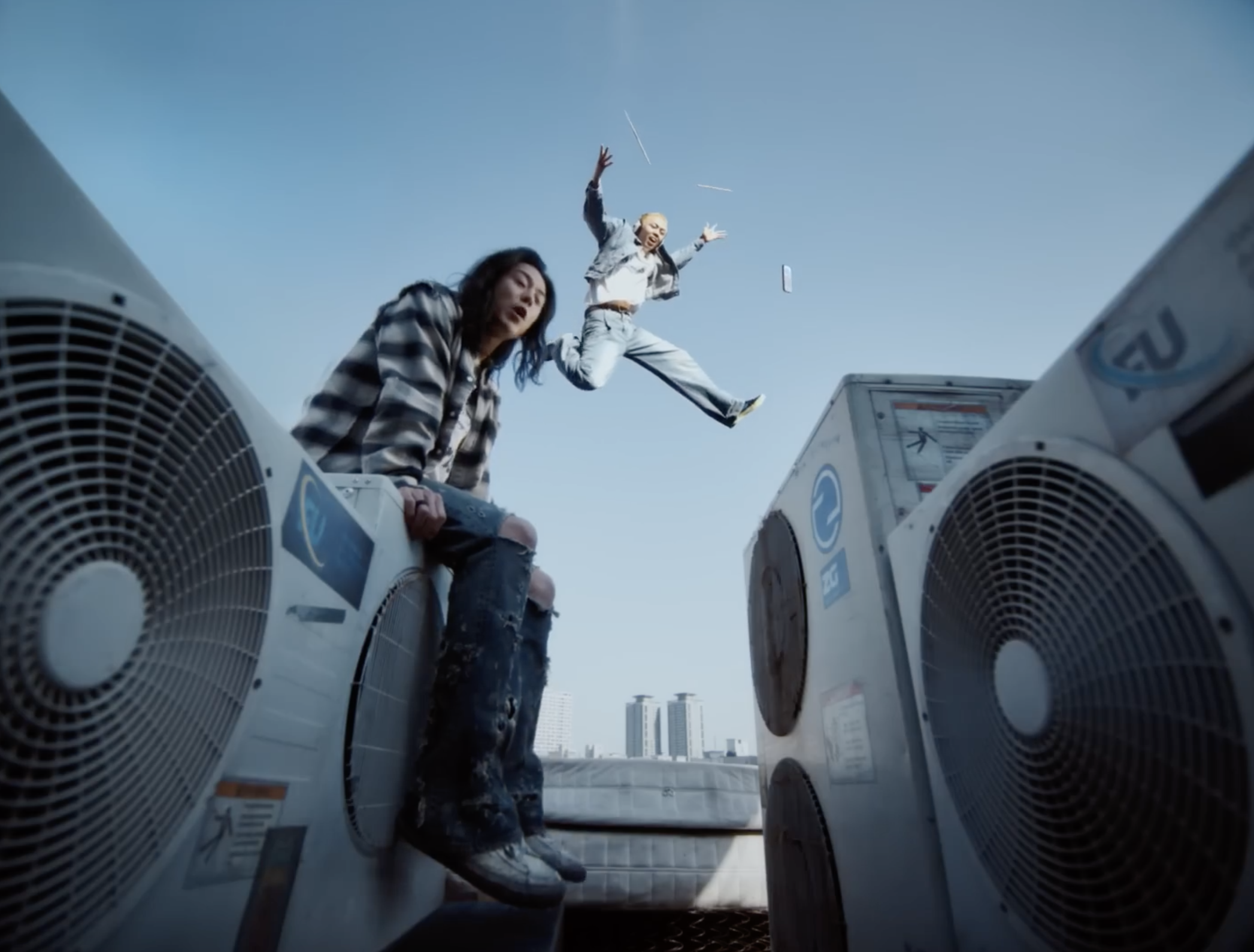 Deux personnes sur un toit avec des climatiseurs, l'une assise et l'autre sautant dans les airs, ciel bleu en arrière-plan.