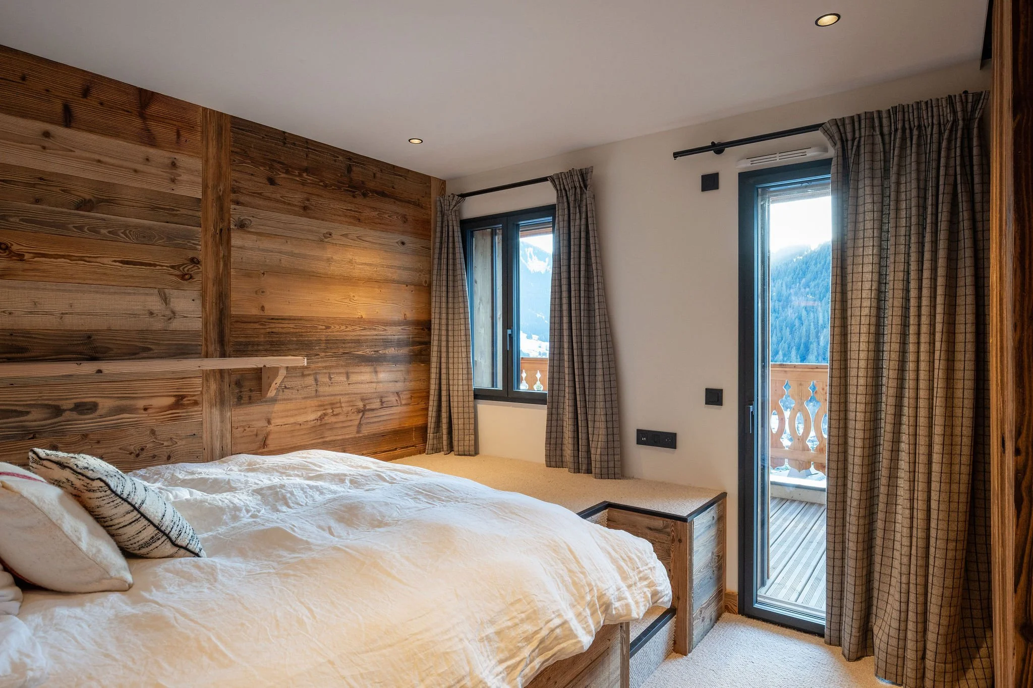 Chambre avec lit en bois, fenêtre et porte donnant sur un balcon en bois, vue montagne, rideaux à carreaux, murs en bois et blanc.