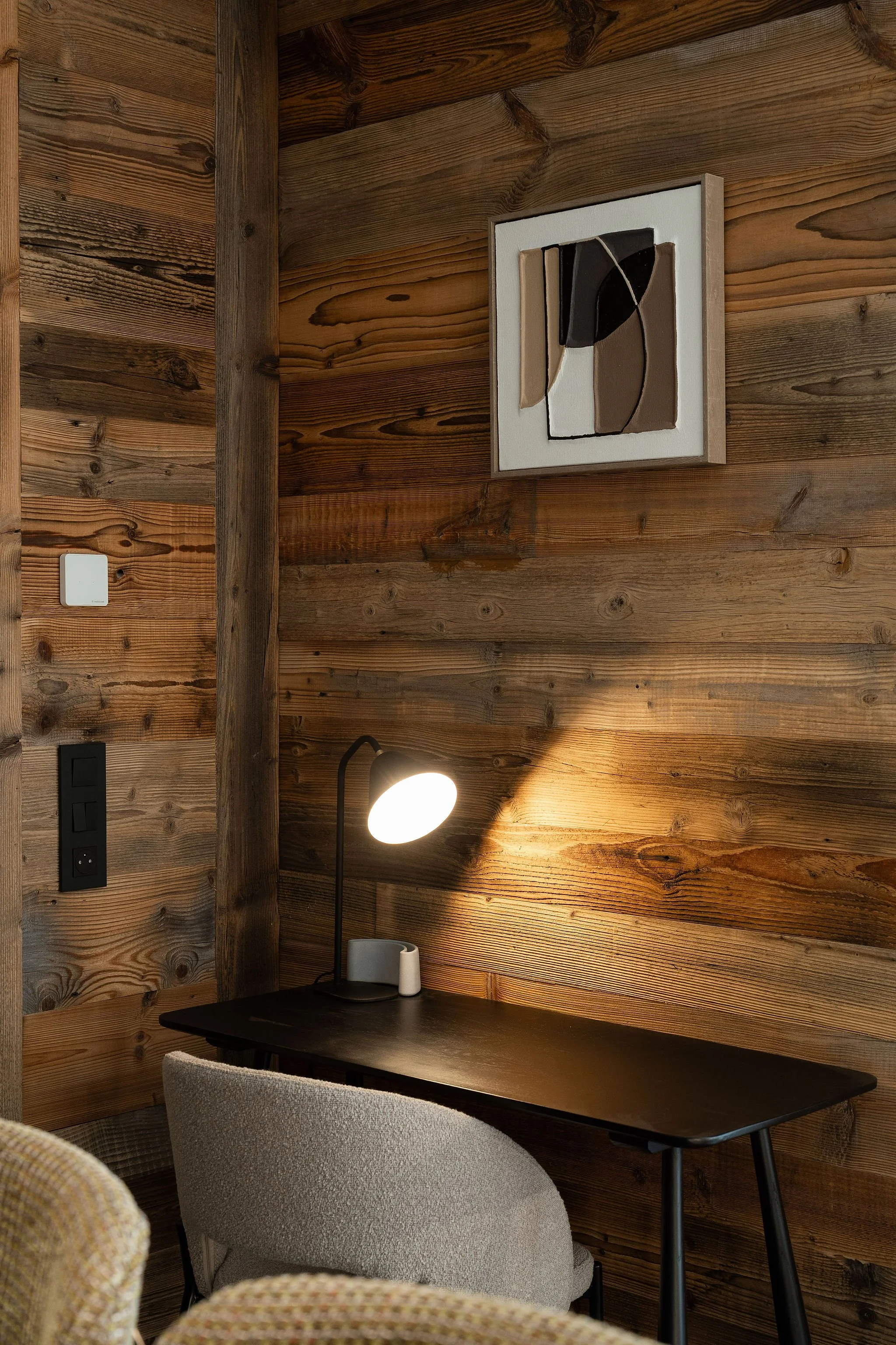 Un bureau avec une table noire, une lampe de bureau allumée, et deux chaises, sur un mur en bois avec un tableau abstrait.