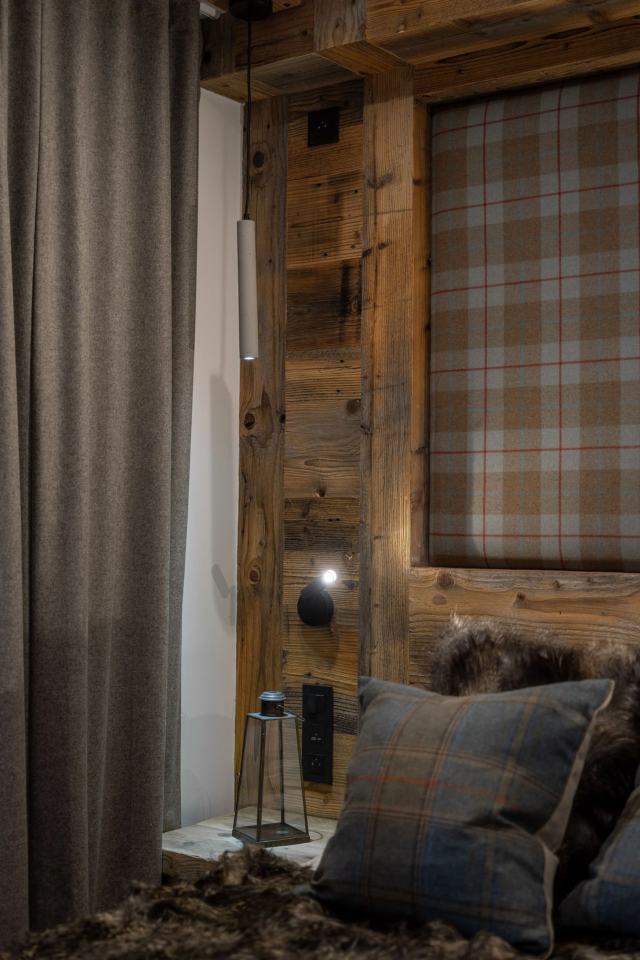 Intérieur d'une chambre avec murs en bois et décoration rustique, contenant un lit avec coussins à carreaux, une lampe de chevet et des prises électriques.