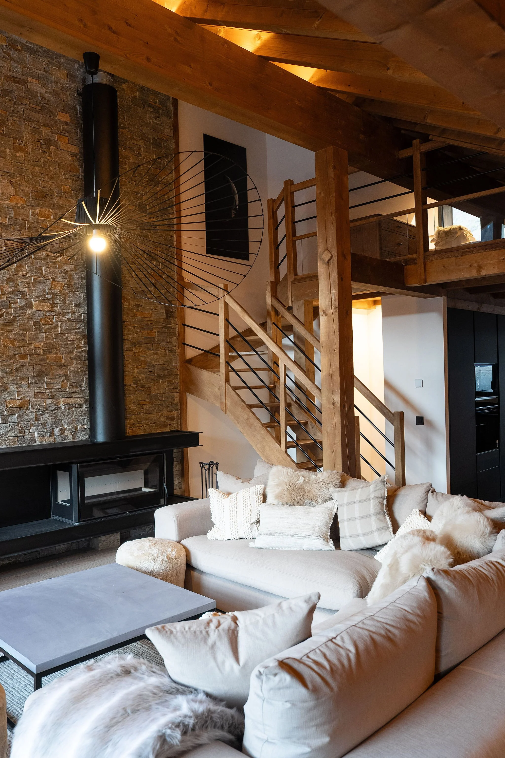 Salon intérieur d'une maison avec canapé beige, plusieurs coussins et tapis en fourrure, escalier en bois menant à une mezzanine, mur en pierre, cheminée noire, décoration moderne dans un style chalet.