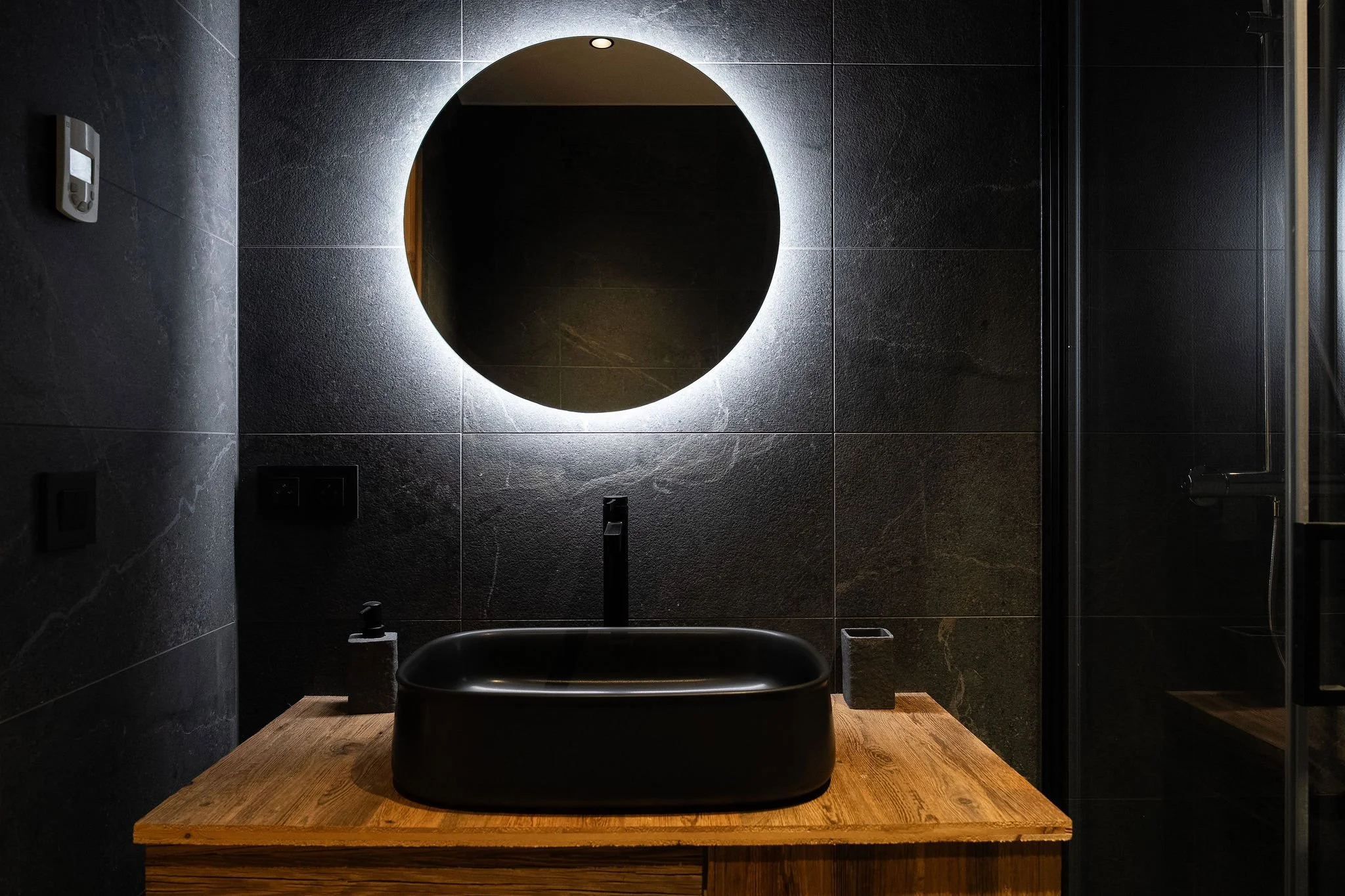 Salle de bain moderne avec un miroir rond éclairé, un lavabo noir sur un meuble en bois, murs en carrelage noir, et une cabine de douche en verre.