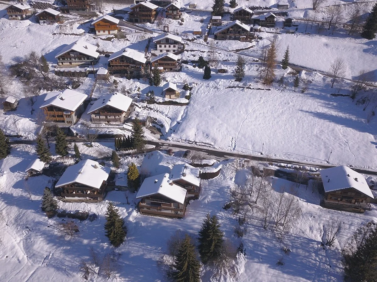 Vue aérienne d'un village alpin enneigé avec des chalets en bois, des arbres et des chemins enneigés.