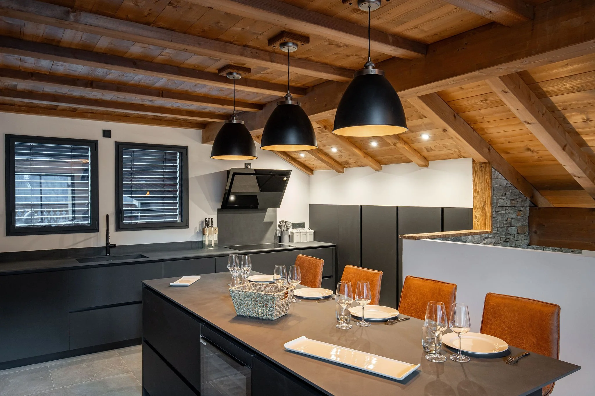 Cuisine moderne avec plafond en bois, trois luminaires noirs suspendus, table avec assiettes et verres, chaises en cuir orange.