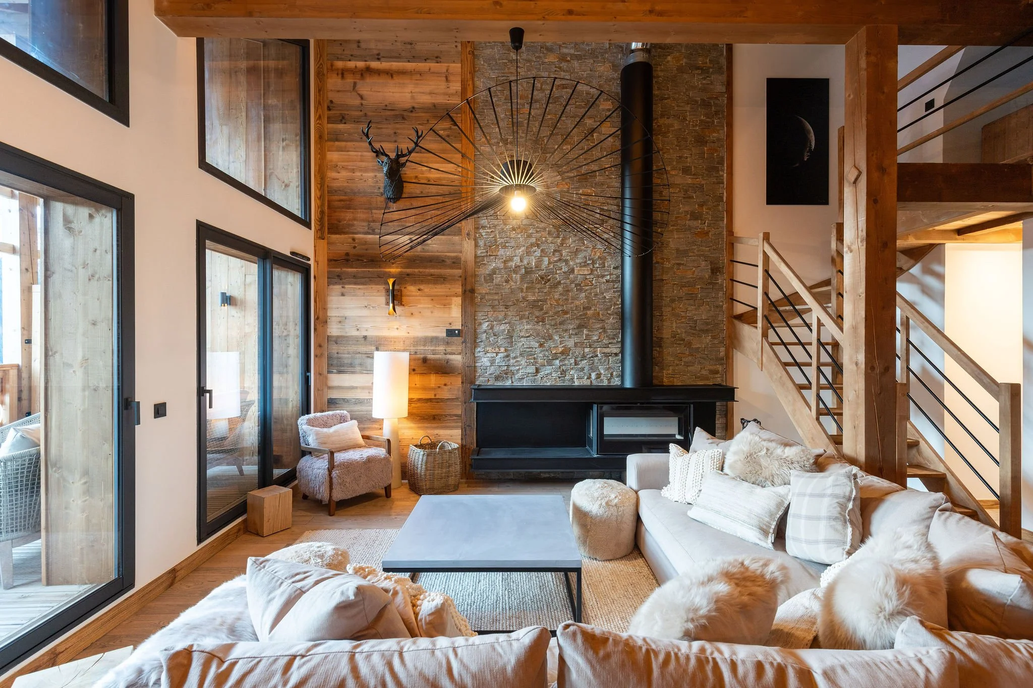 Salon de style montagne avec poêle noir, mur en pierre et bois, canapés en tissu beige avec coussins. Espace lumineux avec grandes fenêtres, escalier en bois et métal, décoration avec une tête de cerf et une œuvre d'art lunaire.