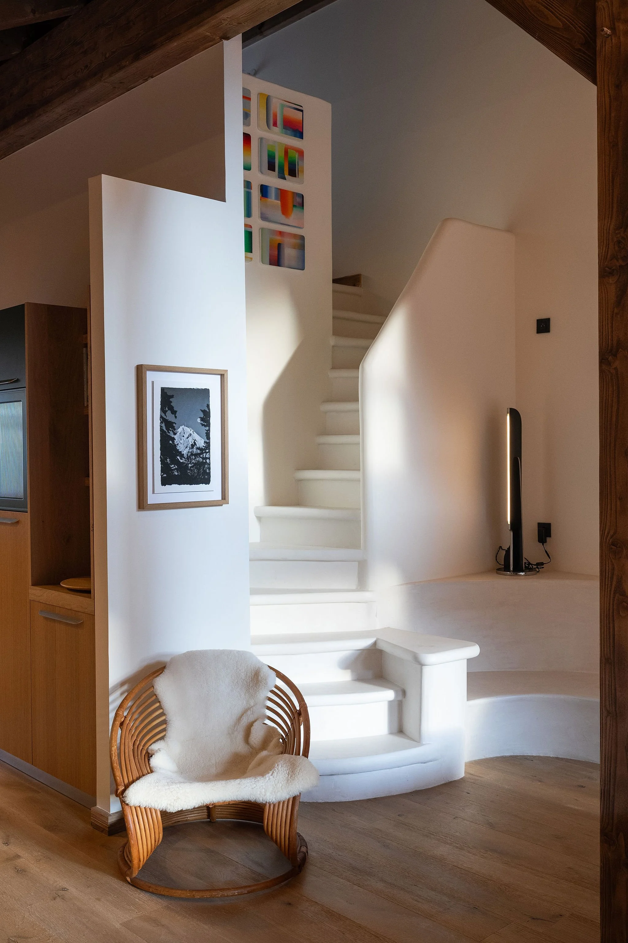 Intérieur d'une maison avec un coin fauteuil en bois avec une couverture blanche, un mur blanc avec une photo noire et blanche, un escalier blanc en colimaçon, une lampe noire moderne, et des œuvres d'art colorées en haut.