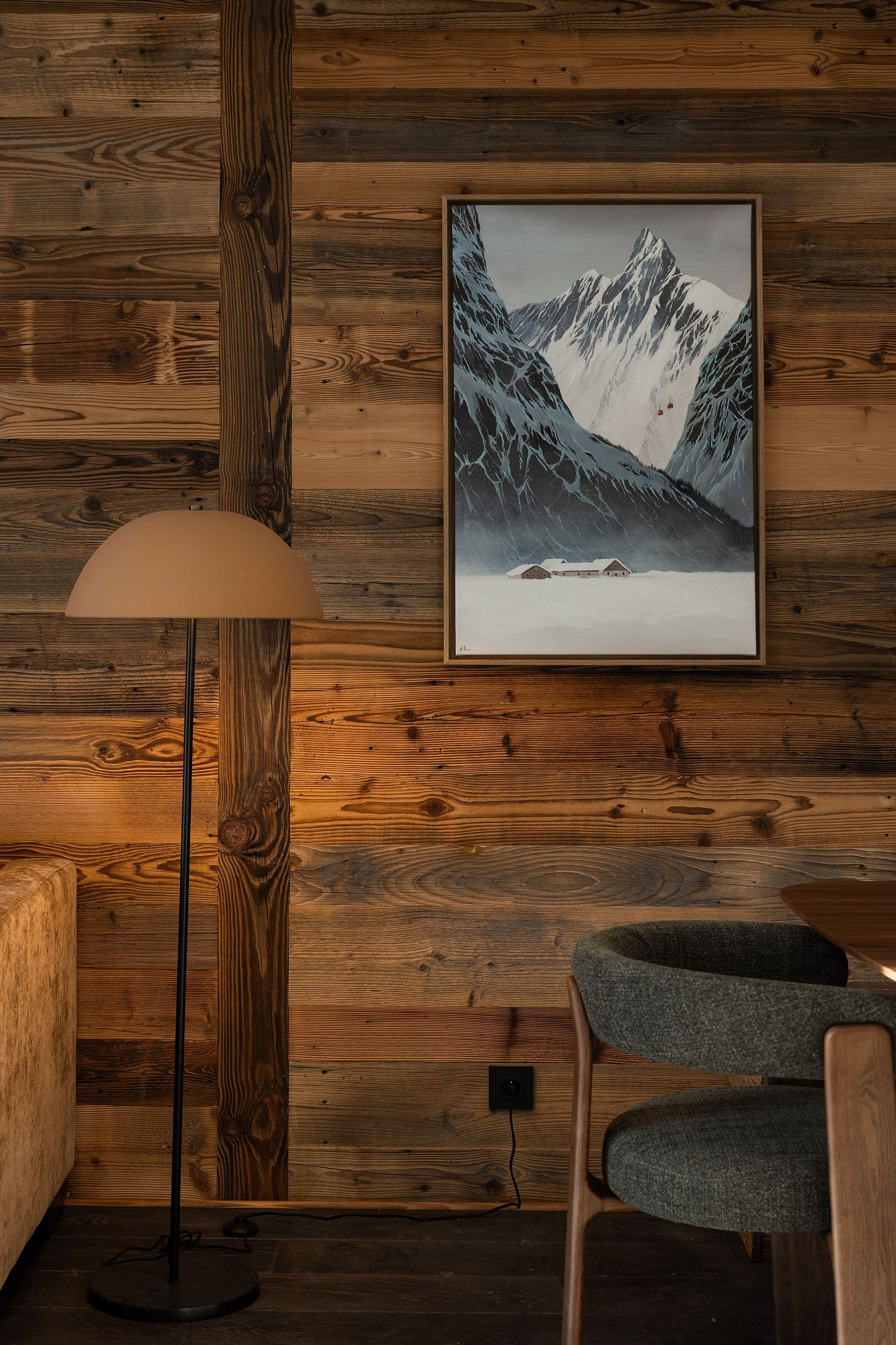 Intérieur avec mur en bois, tableau de montagnes enneigées, lampe de sol beige, fauteuil en tissu gris et un coin de table en bois.