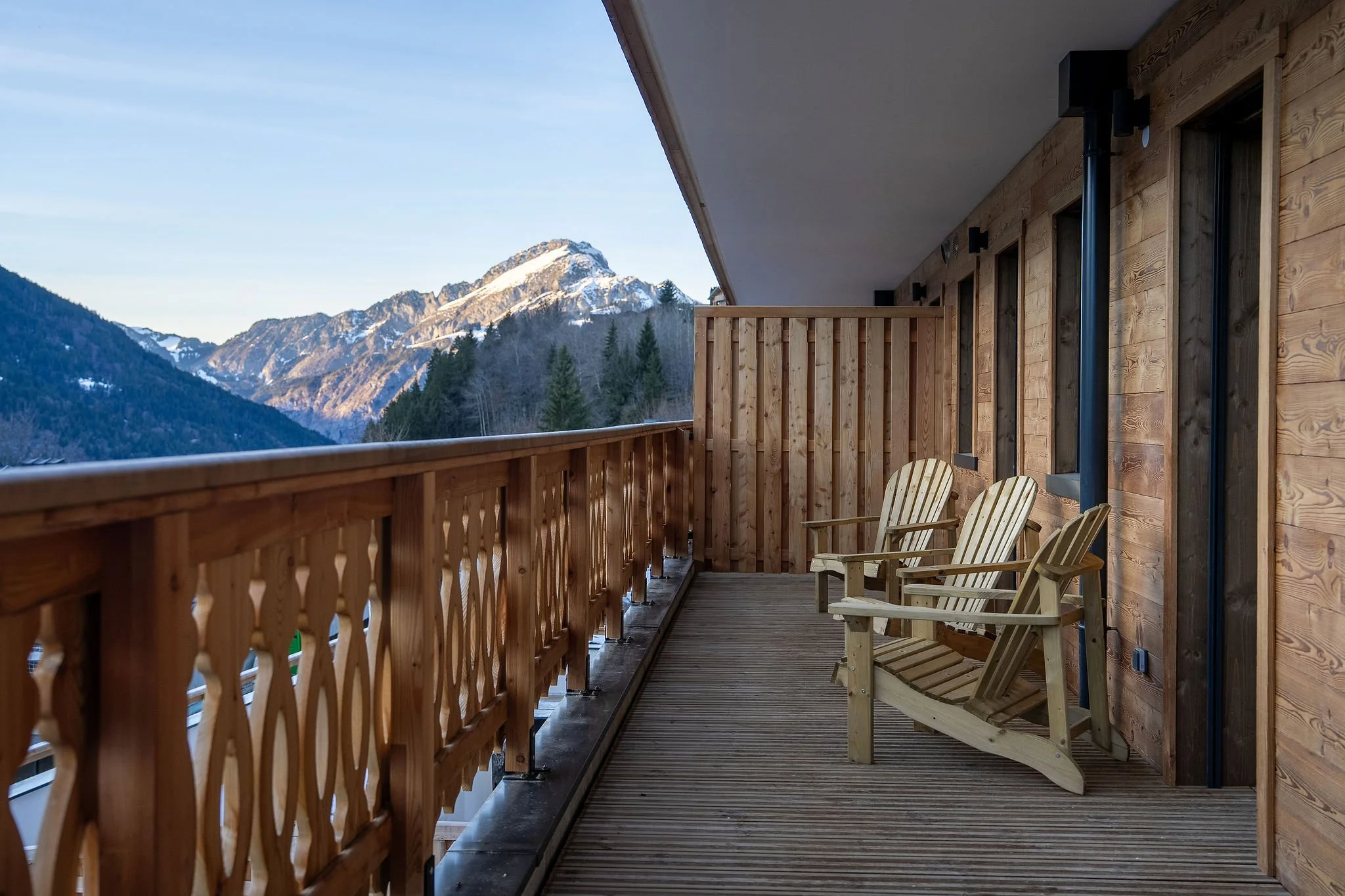 Balcon en bois avec deux chaises en bois, vue sur des montagnes enneigées et une forêt.