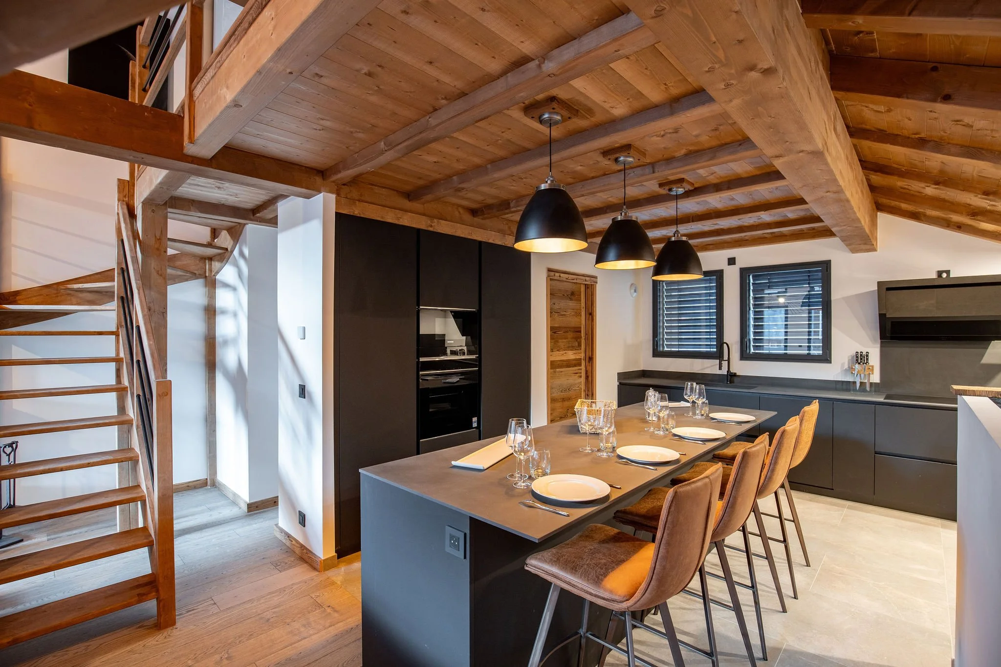 Cuisine moderne avec plafond en bois, comptoir noir, grands fenêtres avec volets noirs, îlot central avec chaises en tissu marron, décor minimaliste, lampe suspendue noire.