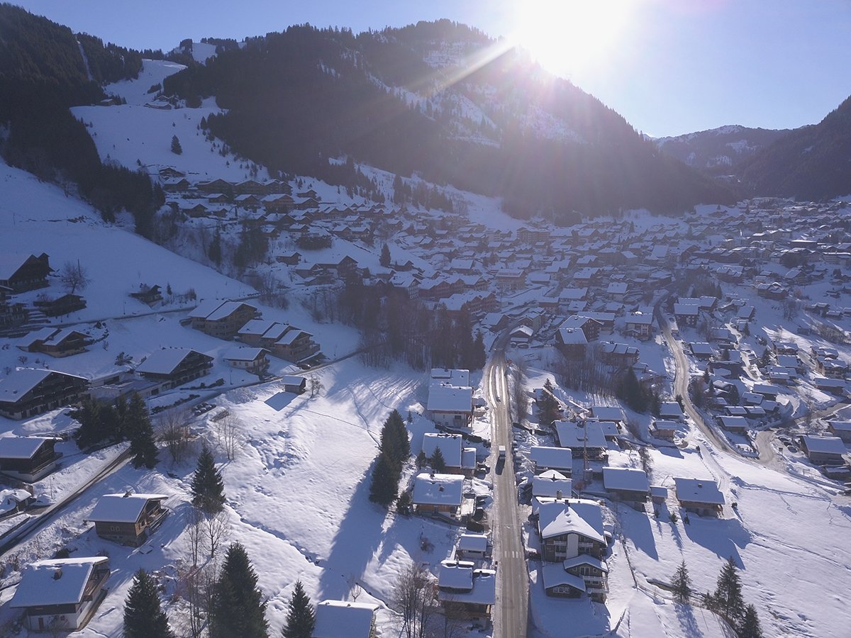 Vallée de montagne enneigée avec des chalets et une route principale. Le soleil brille au-dessus des montagnes.