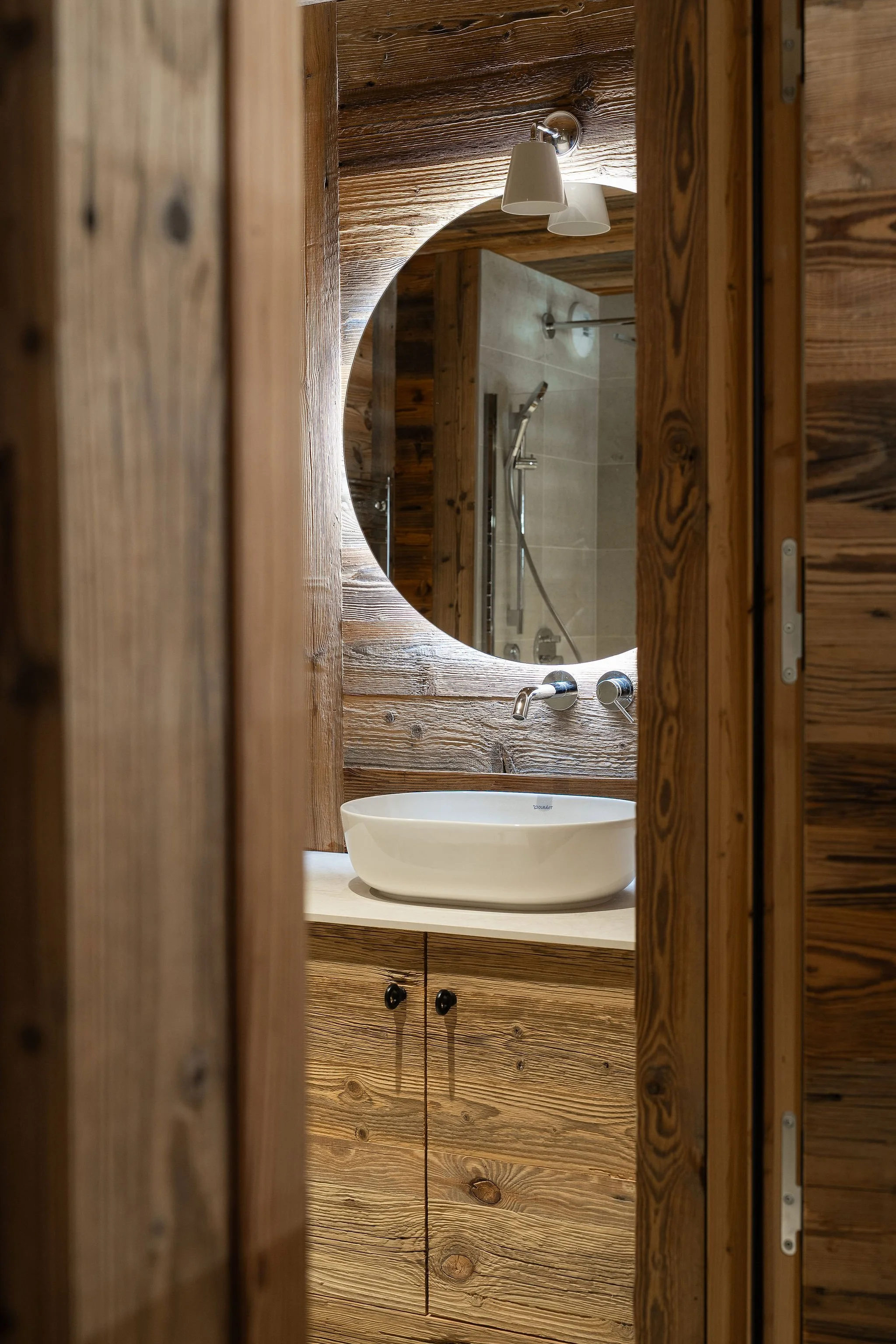 Vue d'un lavabo blanc rond en céramique au-dessus d'un meuble en bois, avec un miroir rond reflétant une douche avec une paroi en verre et une pomme de douche, dans une salle de bain en bois.