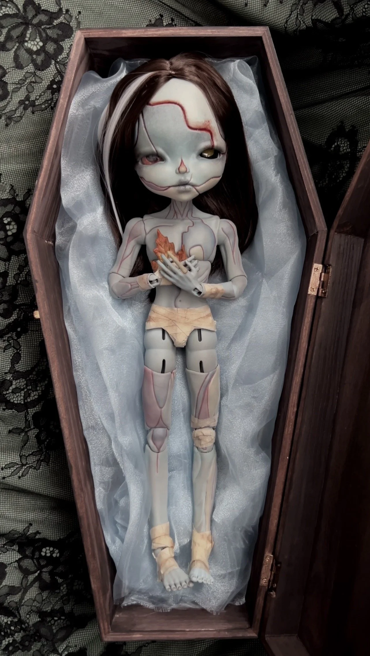 A humanoid doll with a skeletal and robotic appearance, lying in a wooden box with satin lining. The doll has a pale face with painted red and black markings, glowing eyes, and long brown and white hair. Its body is designed with mechanical and skele