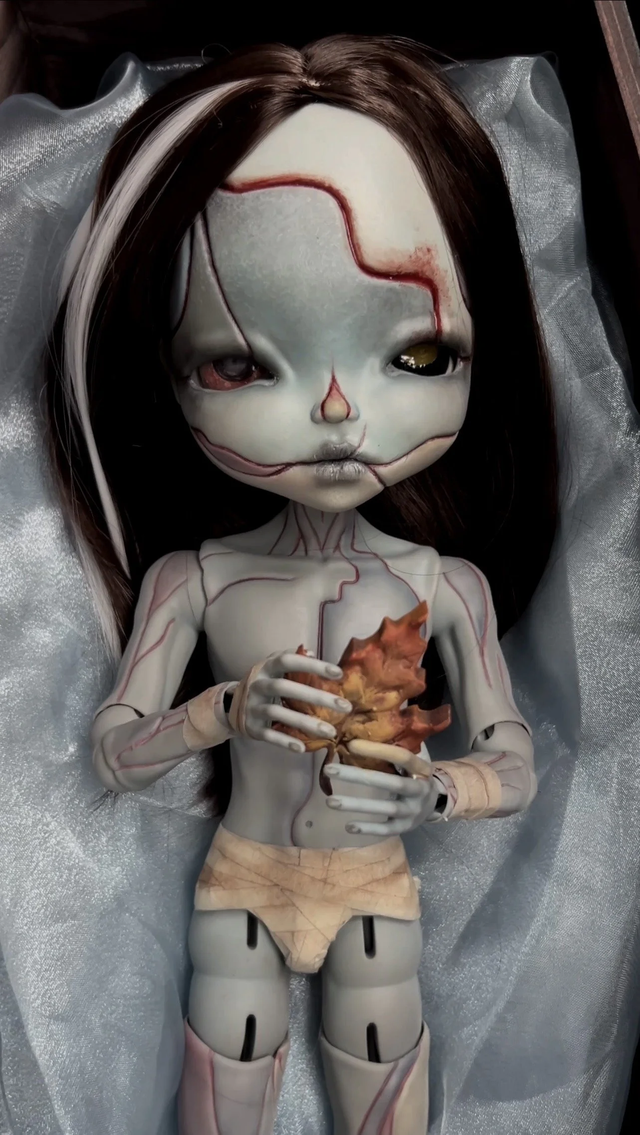A doll with a pale, cracked face and dark brown hair lies on a satin surface, holding a realistic, anatomical organ in its hands.