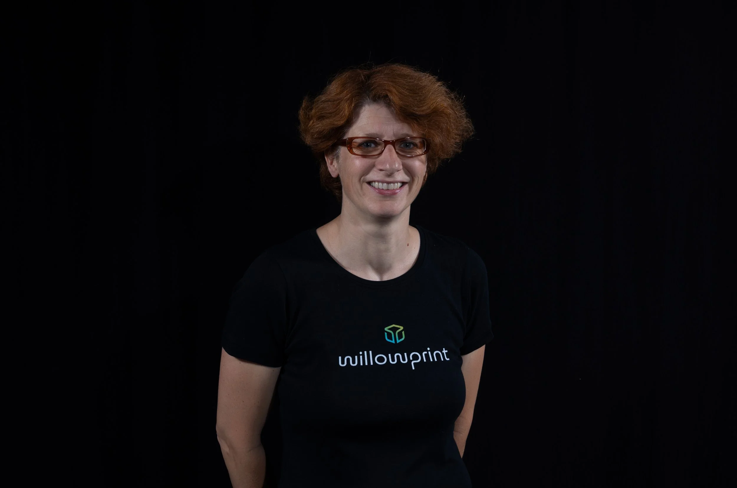 A woman with red hair smiling, wearing a black turtleneck and a black t-shirt with a logo and the text 'willowprint' against a dark gray background.
