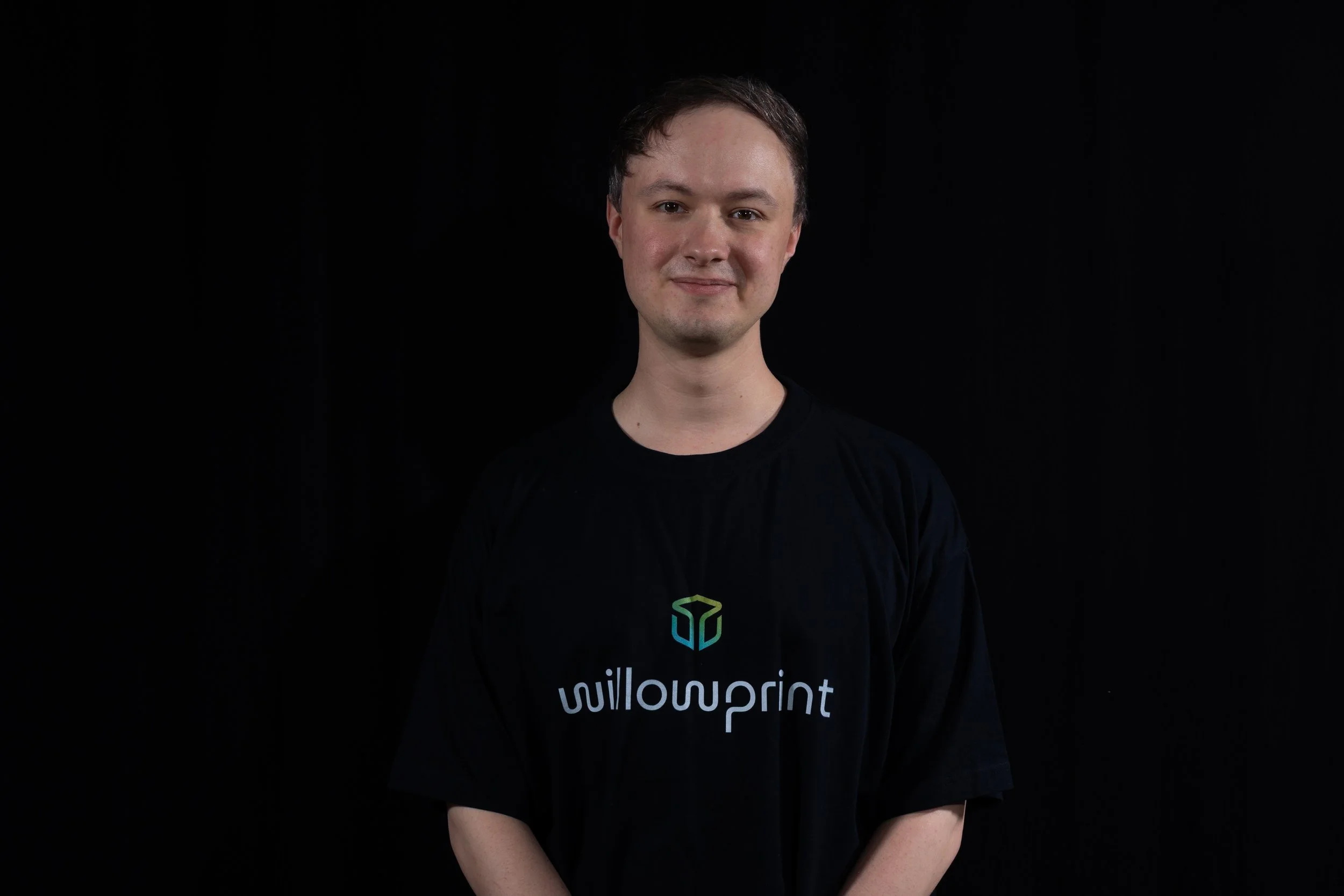 A woman with shoulder-length brown hair wearing a black t-shirt with willowprint logo, standing against a dark gray background.