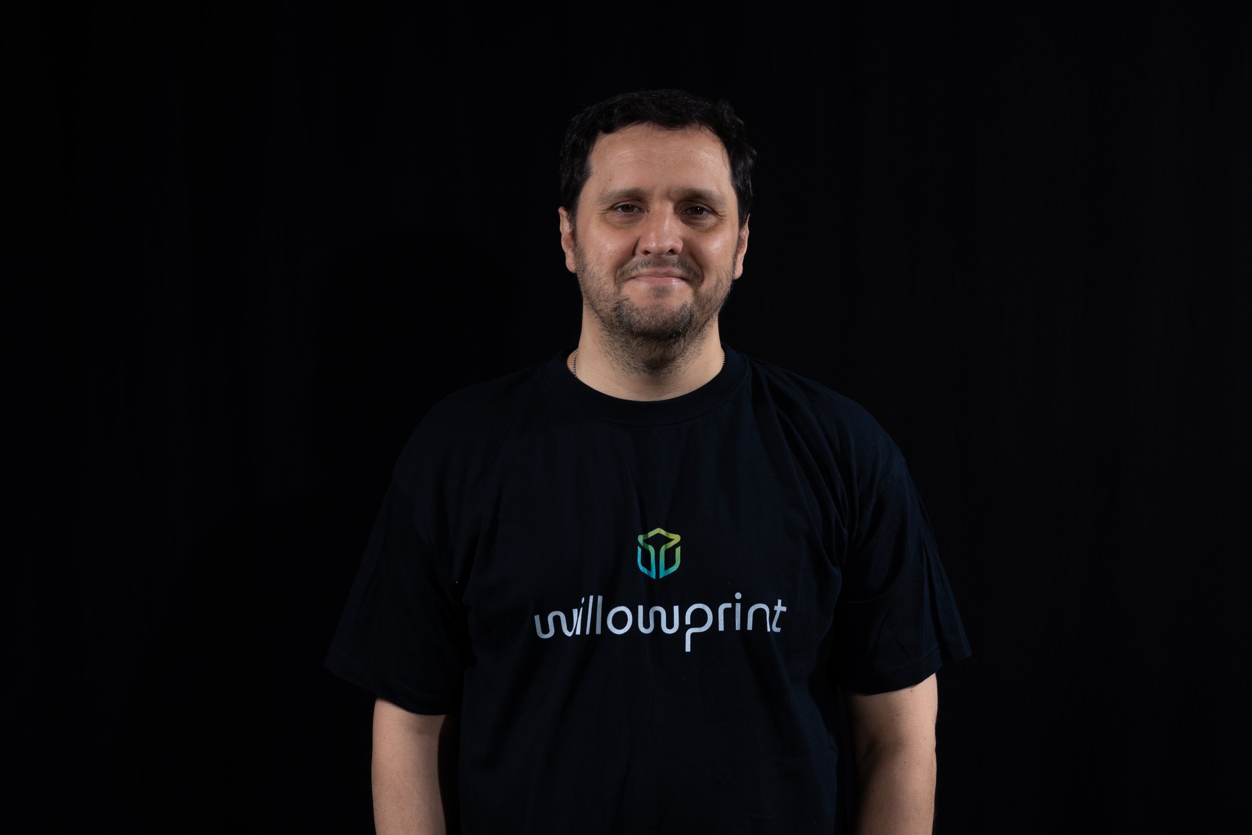 A man with dark hair and a slight smile wearing a black t-shirt with the 'willowprint' logo standing against a dark gray background.