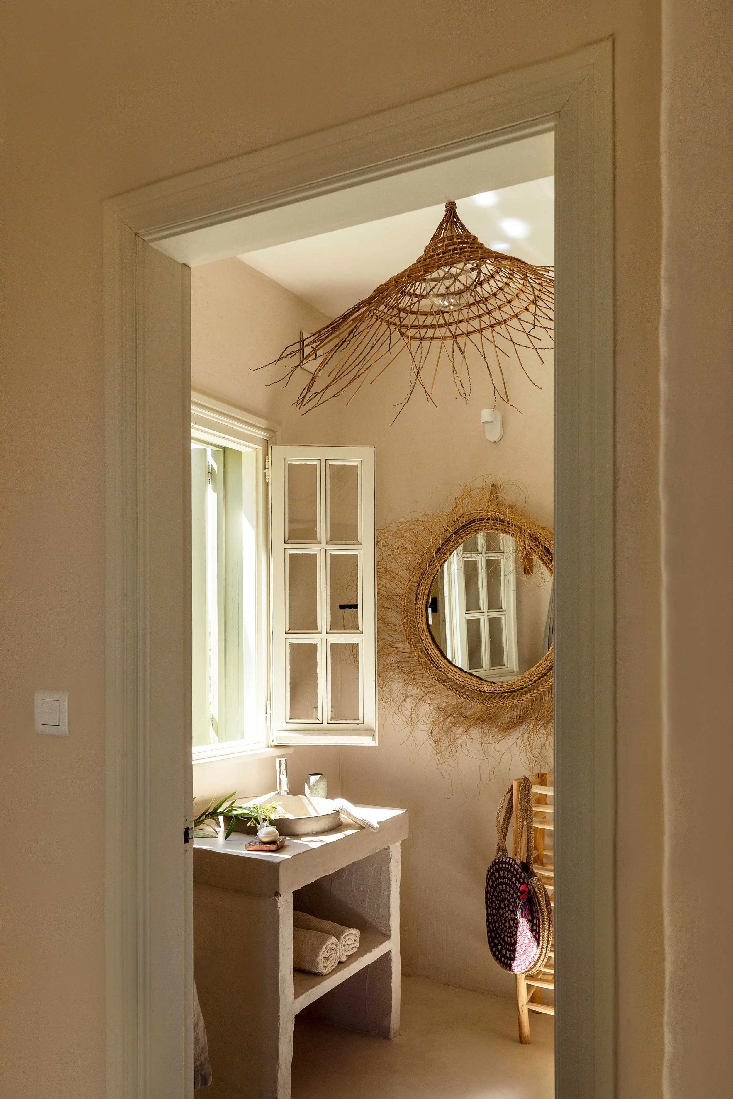 A cozy bathroom with a open window, a round mirror with a woven frame, and a hanging wicker light fixture.