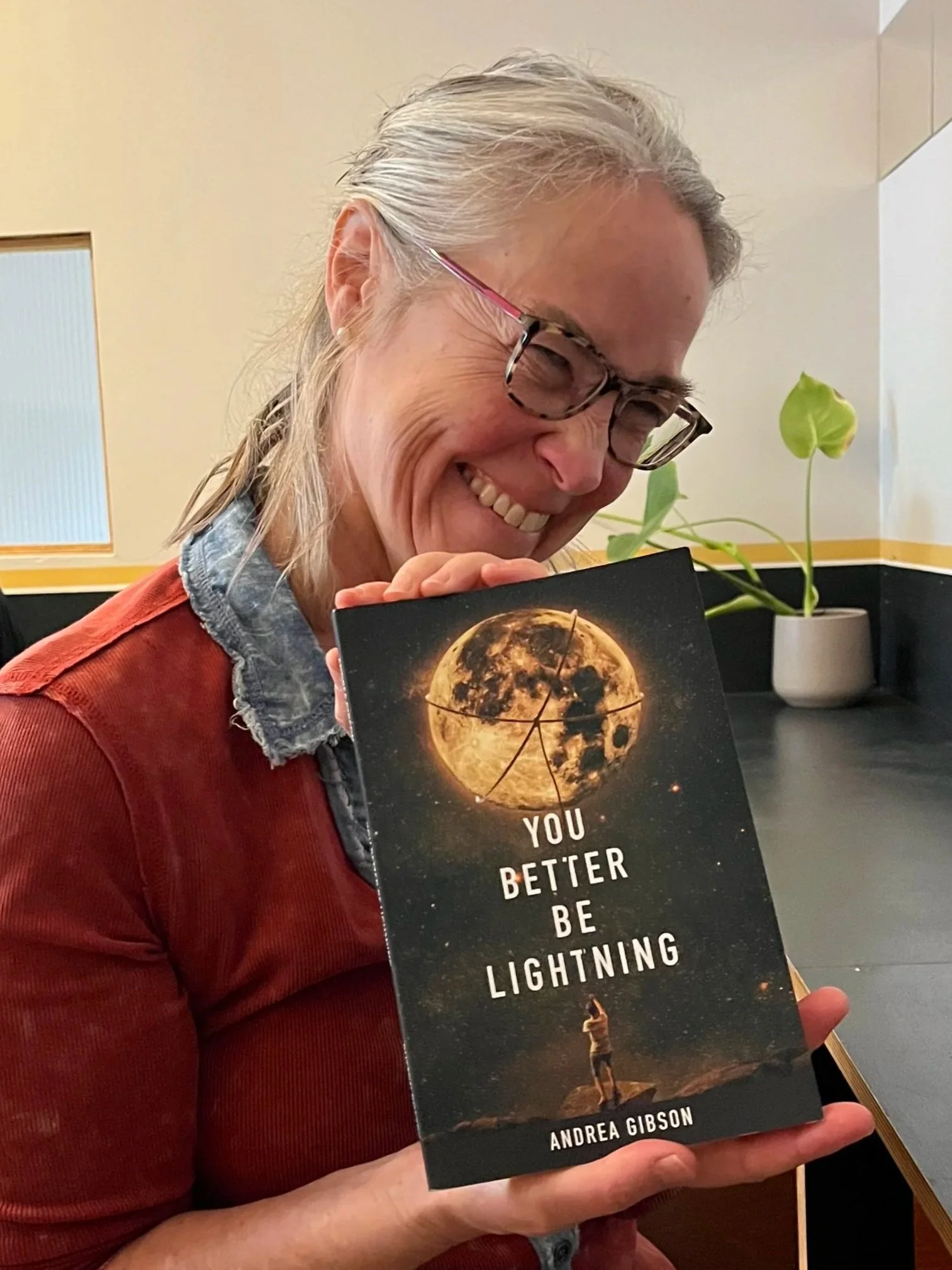 A smiling woman with glasses holding a book titled 'You Better Be Lightning' by Andrea Gibson, with a cosmic background featuring the moon and stars and a person standing on a rock.