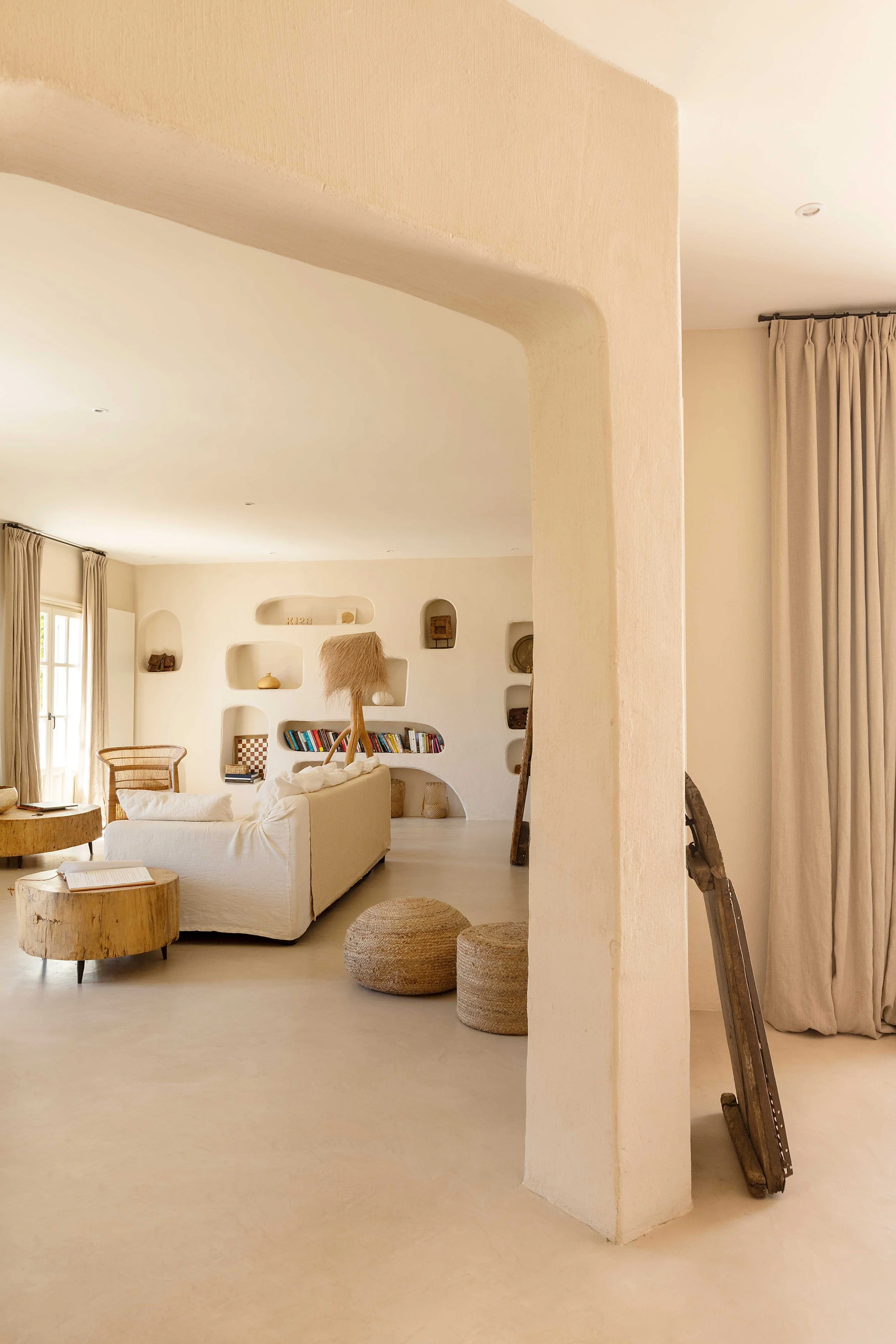 A cozy, neutral-toned living room with built-in wall shelves holding decorative items, a white sofa, round ottomans, and rustic furniture, with curtains on windows and natural lighting.