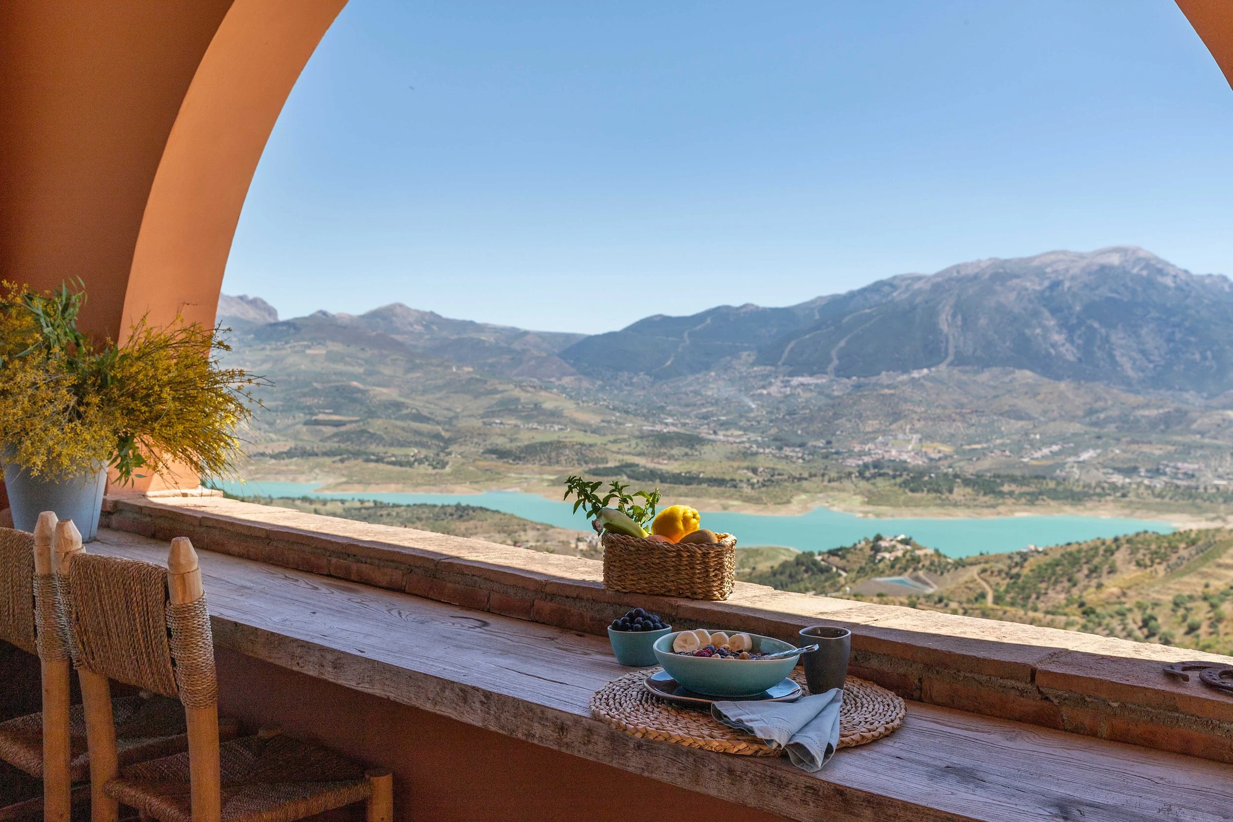 A scenic balcony view of a mountainous landscape with a lake, featuring a wooden table set with a basket of lemons, a bowl of blueberries, bananas, and a black mug, with a small plant on the side.