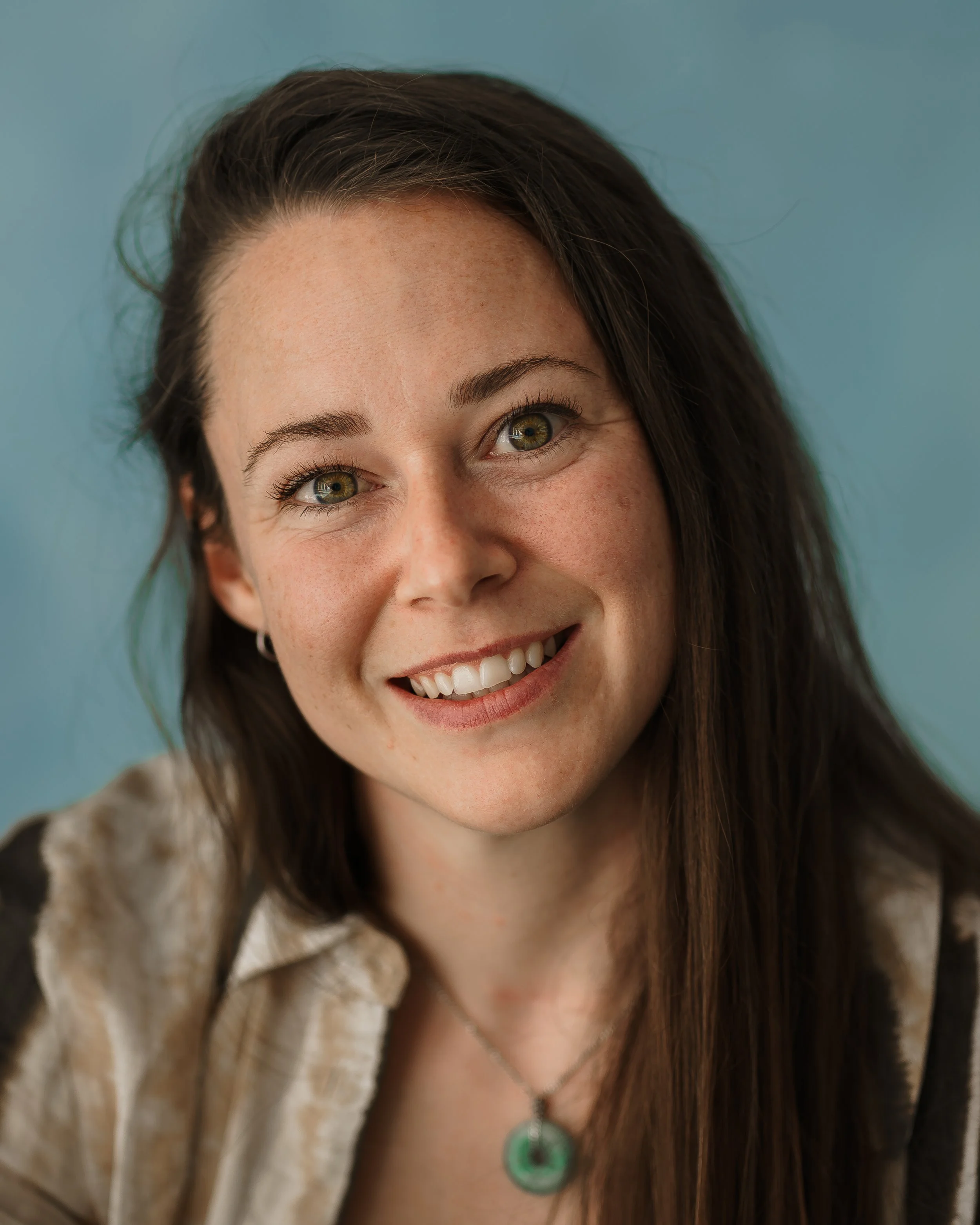 Close-up of a smiling woman with long dark hair, green eyes, wearing a necklace with a green pendant, against a blue background.