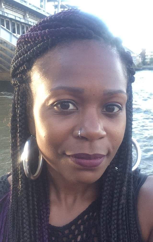 A woman with braided hair, wearing large hoop earrings and a septum nose ring, standing near water with buildings in the background.