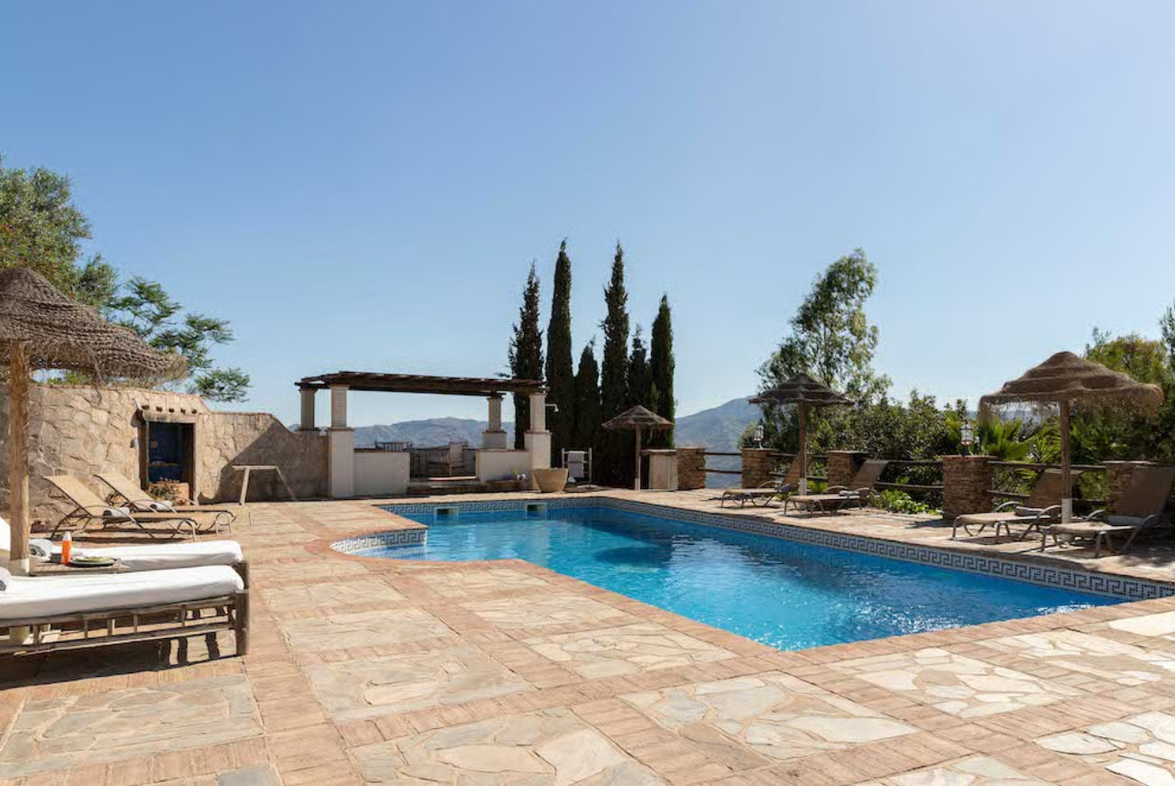 A luxurious outdoor swimming pool area with stone flooring, surrounded by sun loungers with umbrellas, a seating pavilion, greenery, and hills in the background.