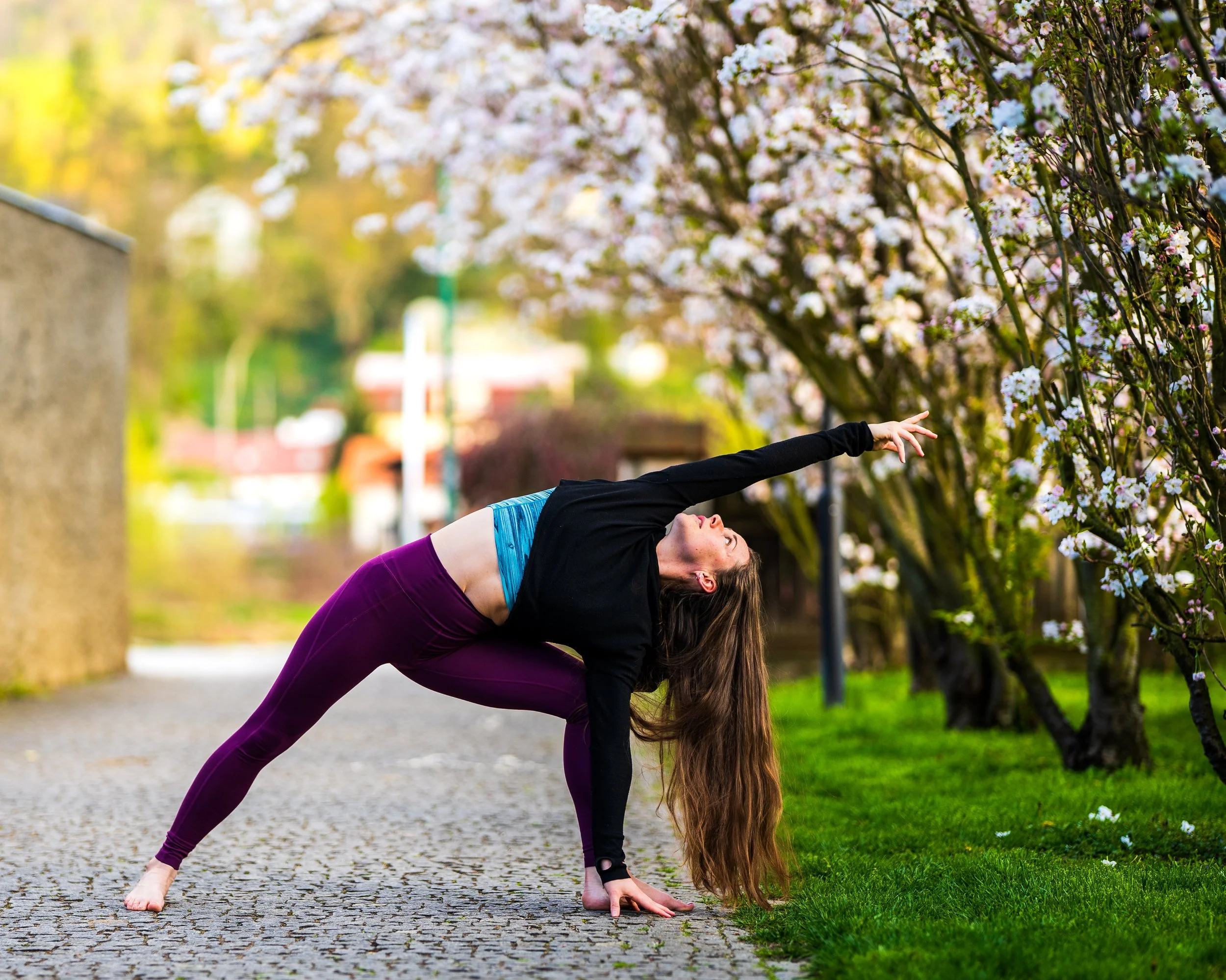 A woman practicing yoga outdoors on a paved path next to blooming trees with pink and white flowers.