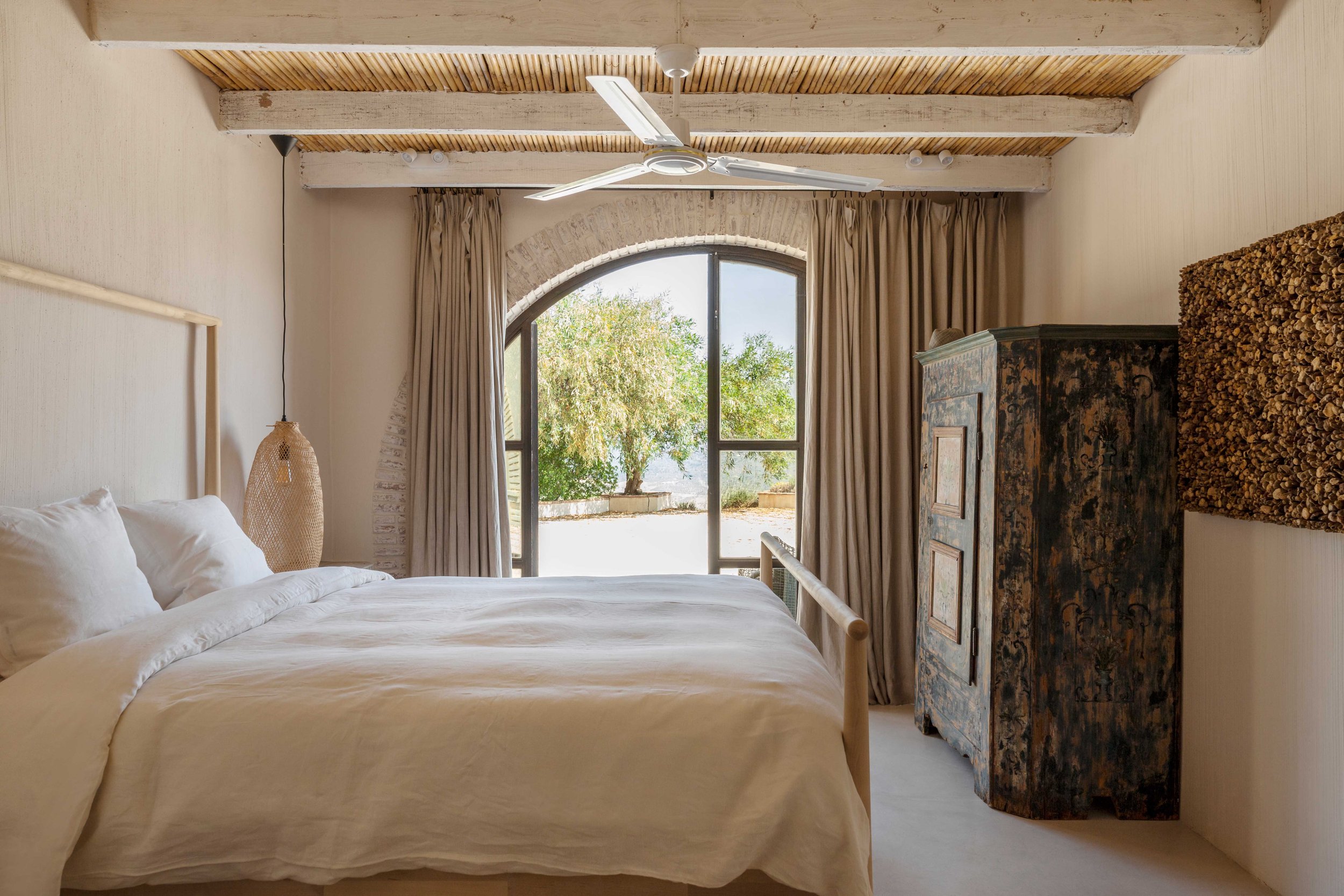 Bedroom with white bed, beige curtains, wooden ceiling beams, ceiling fan, arched window showing trees outside, and vintage-style blue cabinet