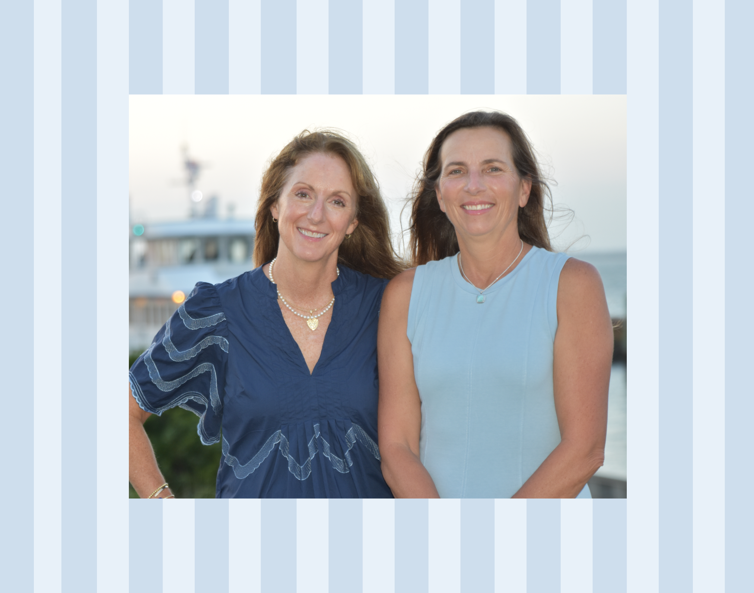 Two smiling women standing outdoors near a marina, with a boat visible in the background. One woman has long brown hair and is wearing a blue dress, the other woman has dark hair and is dressed in a light blue top.