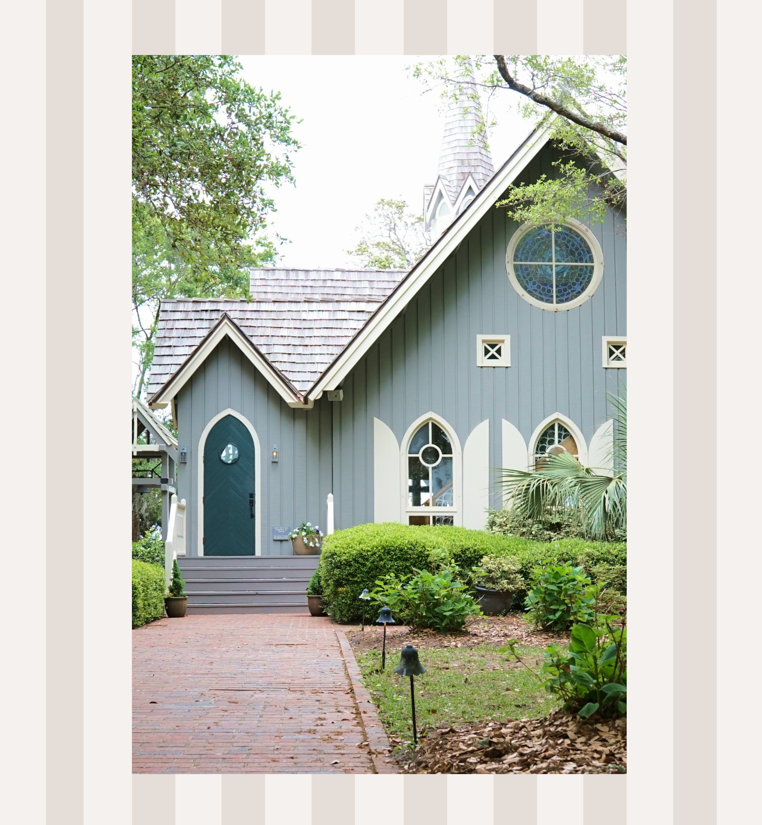 Front view of a church with a circular stained glass window, pointed arch door, grey siding, and shingled steeple, surrounded by trees and bushes.
