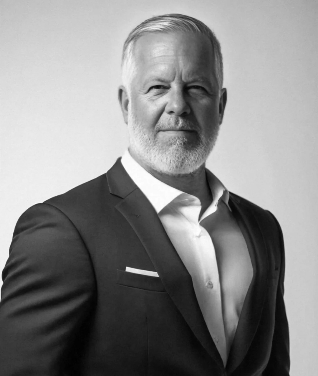 Black and white photo of a man with gray hair and beard in a suit, looking at the camera, standing against a plain background.