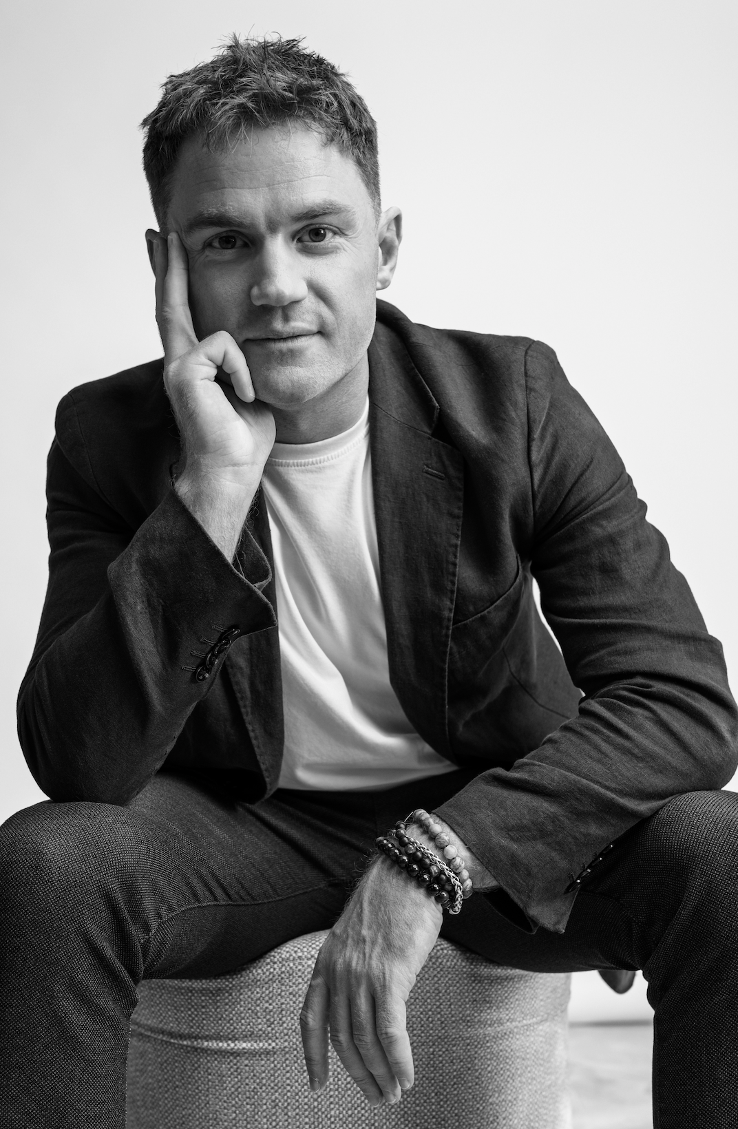 Black and white portrait of a man sitting on a textured stool, with his right hand resting on the side of his face, wearing a dark blazer, a white t-shirt, and multiple beaded bracelets on his left wrist, with a plain background.