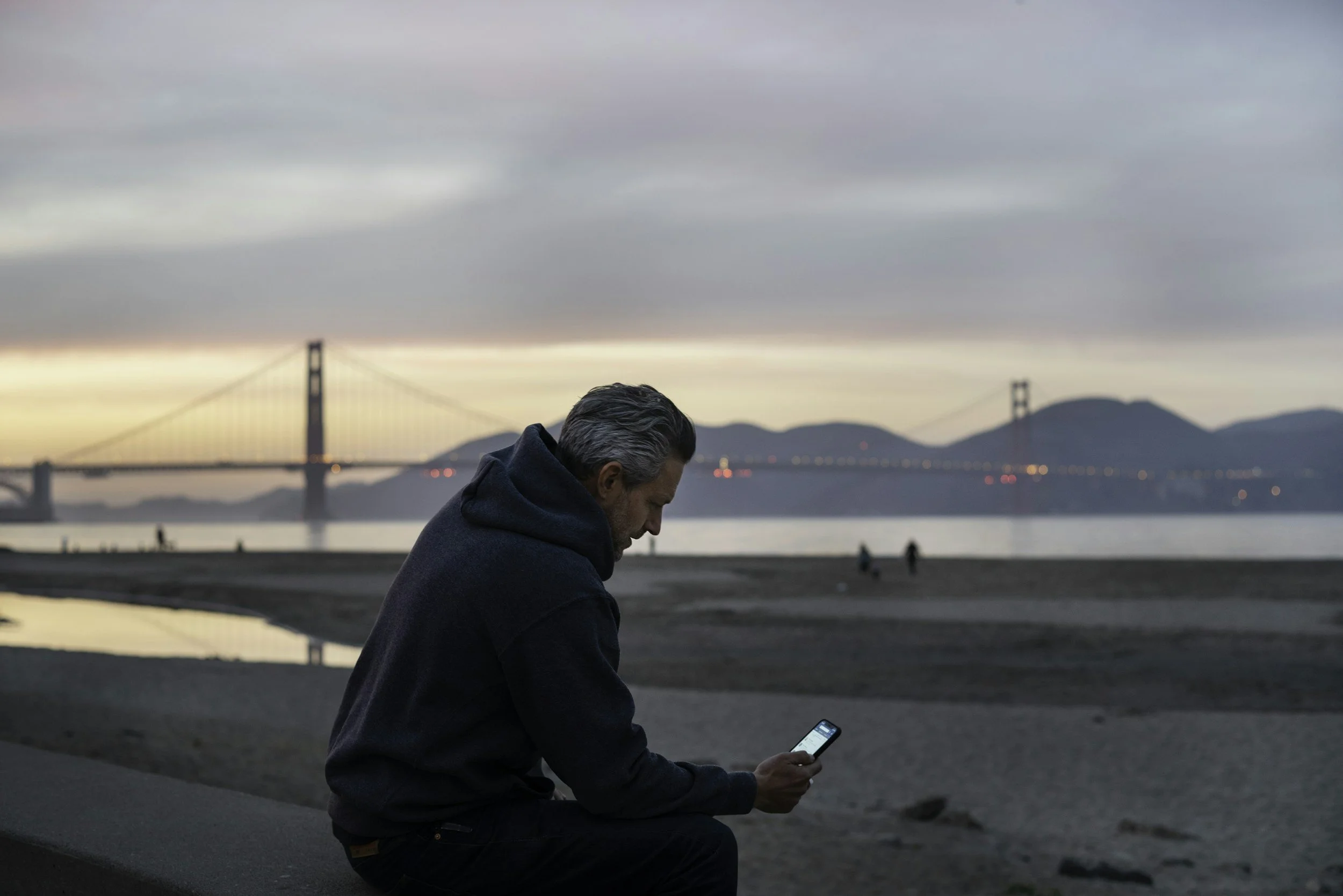 A man in a dark hoodie sitting on a ledge at the beach, looking at his phone, with the Golden Gate Bridge in the background at sunset.