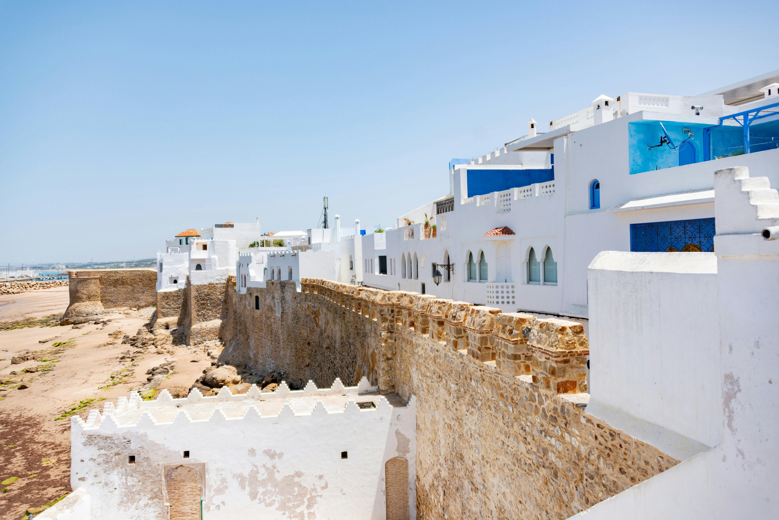 White and blue coastal houses with stone fortifications along a beach under a clear blue sky.