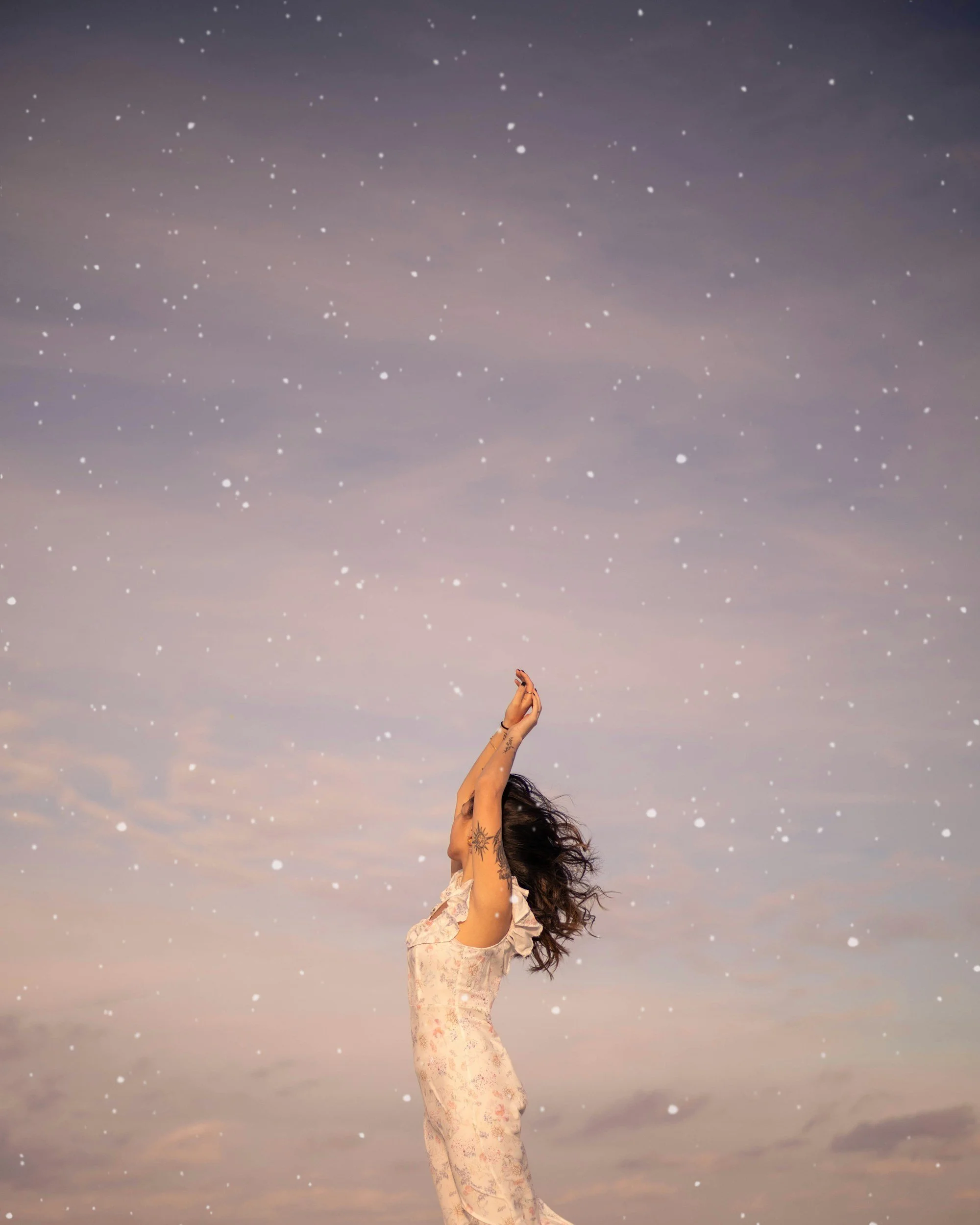 A woman standing outdoors under a sky with clouds and falling snow, stretching her arms upwards because of bold moves coaching for midlife women.